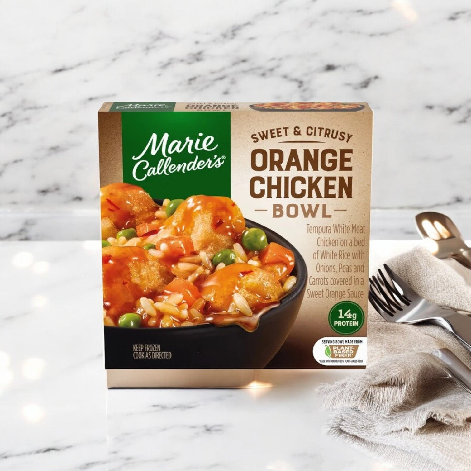 A Smart Ones Marie Callender's Sweet & Citrusy Orange Chicken Bowl, 11 oz., rests on a table. The package showcases the orange chicken with an enticing dish image alongside a fork and napkin, set against a blurred bokeh backdrop.