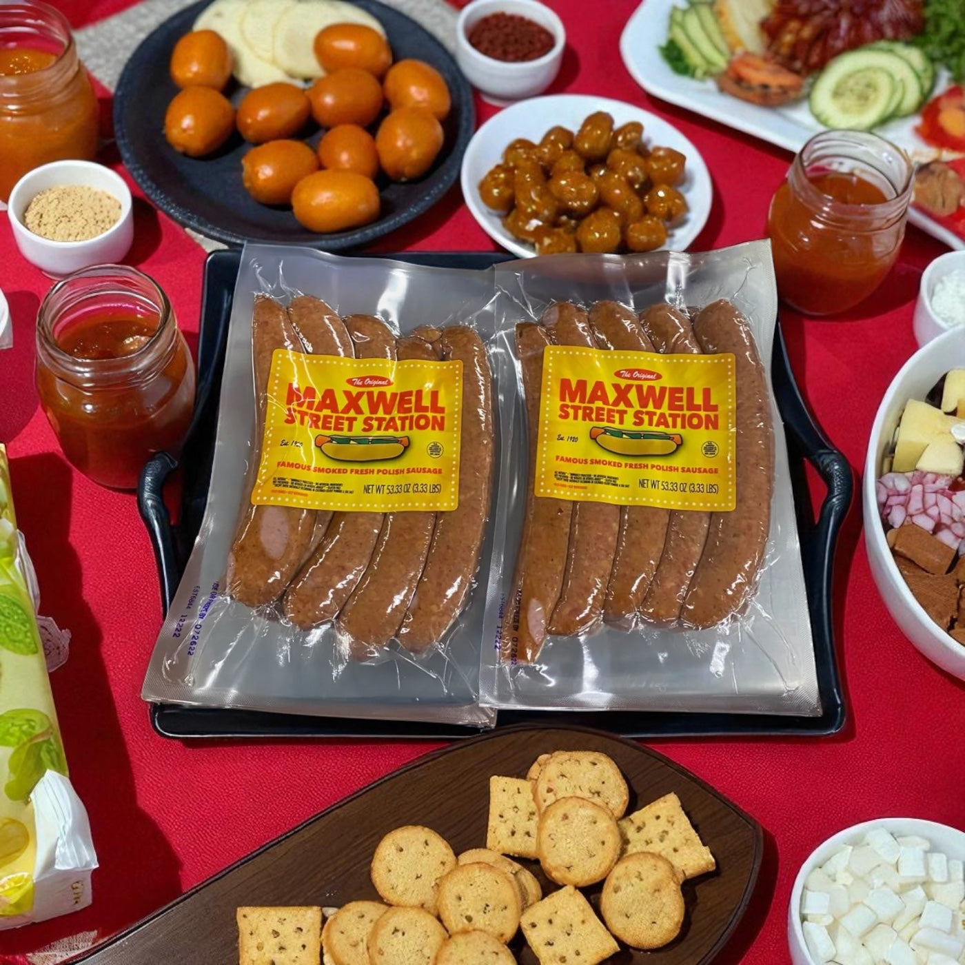 A pack of Vienna Beef Maxwell Street Station Famous Smoked Fresh Polish Sausage (53.33 oz.) is displayed on a tray with crackers, cheese, fruit, sauces, and assorted snacks atop a red tablecloth.