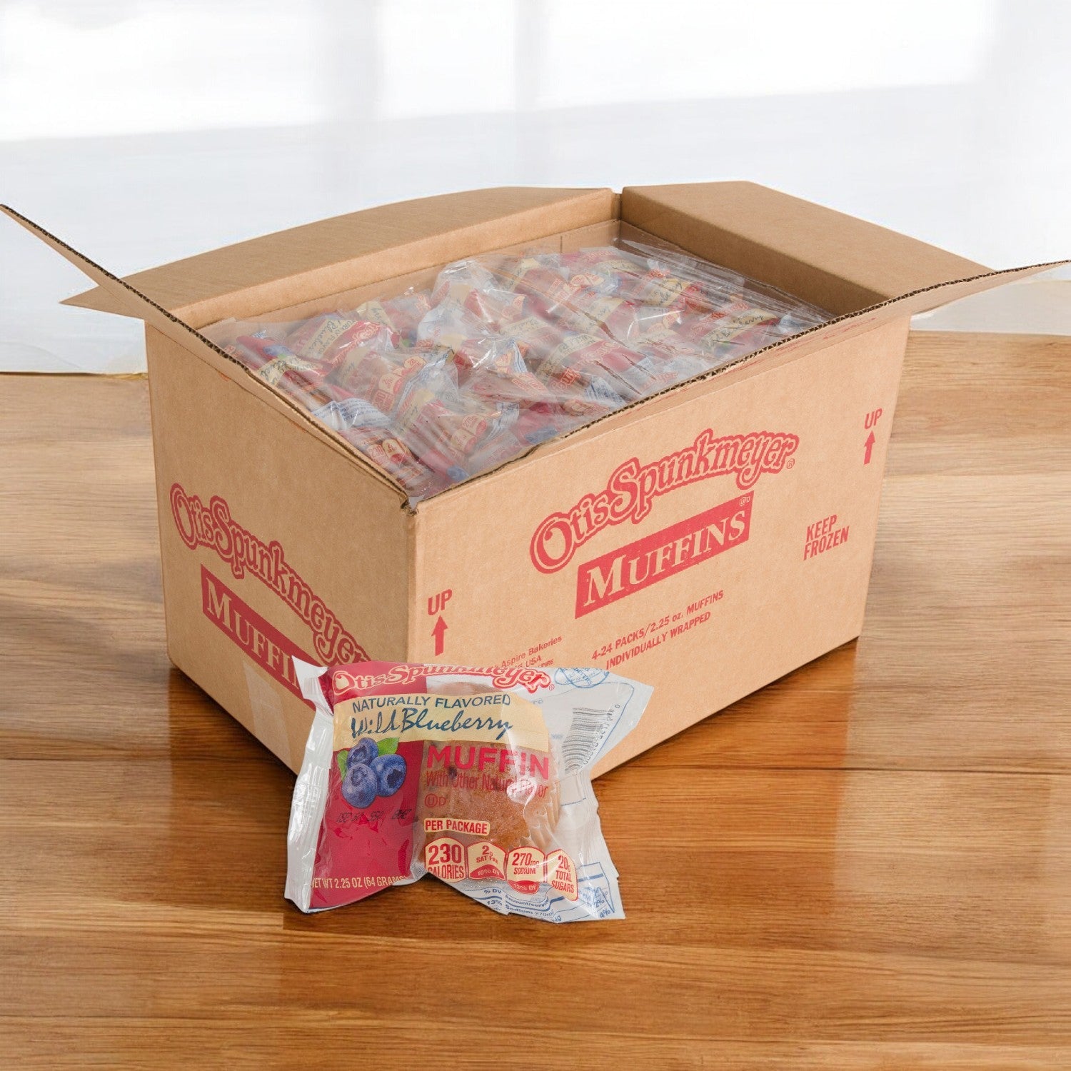 A case of Otis Spunkmeyer Muffin Blueberry Individually Wrapped Frozen (96 count) is on a wooden surface, showcasing one invitingly at the front.