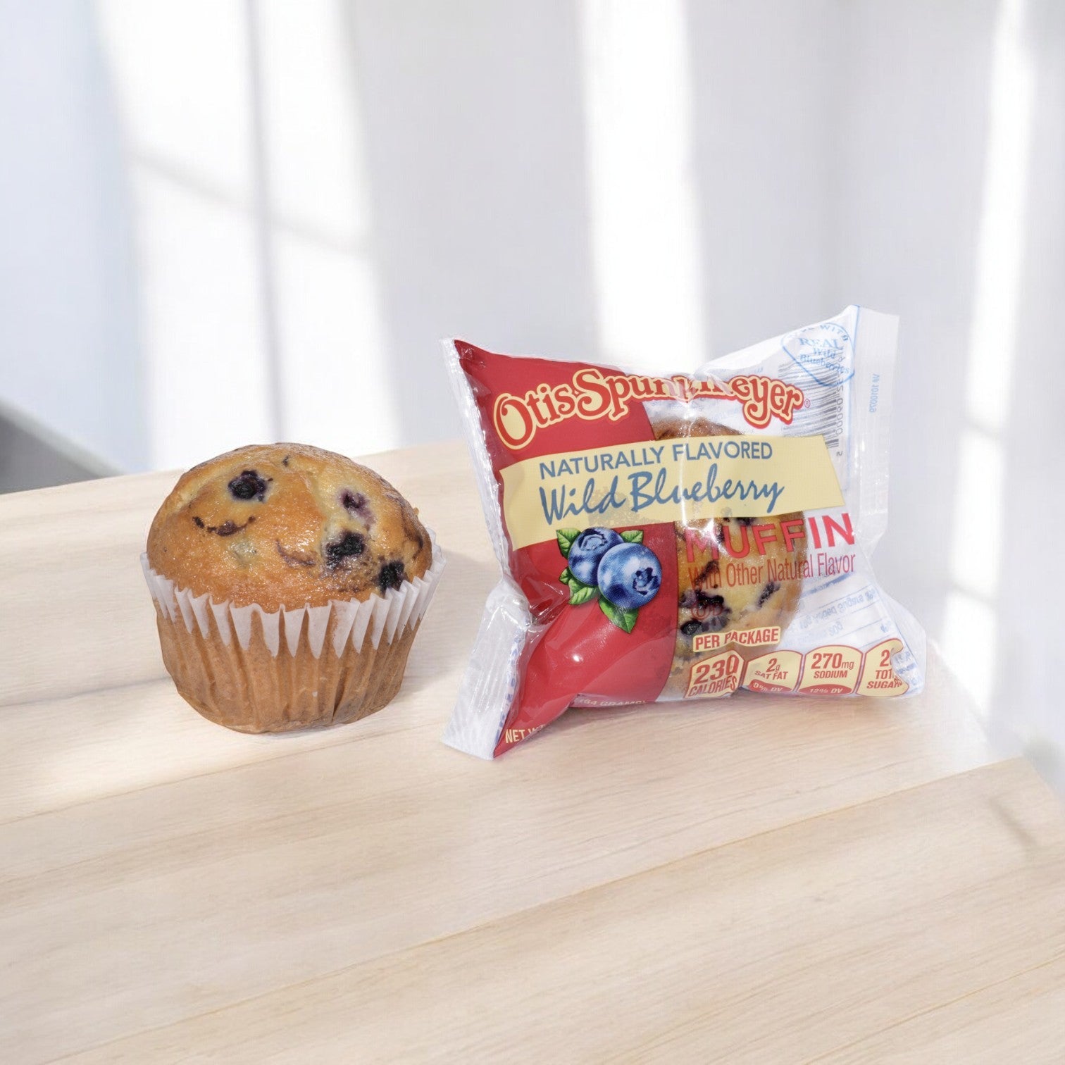A blueberry muffin sits on a wooden surface next to an individually wrapped Otis Spunkmeyer Blueberry Muffin from a frozen case of 96.