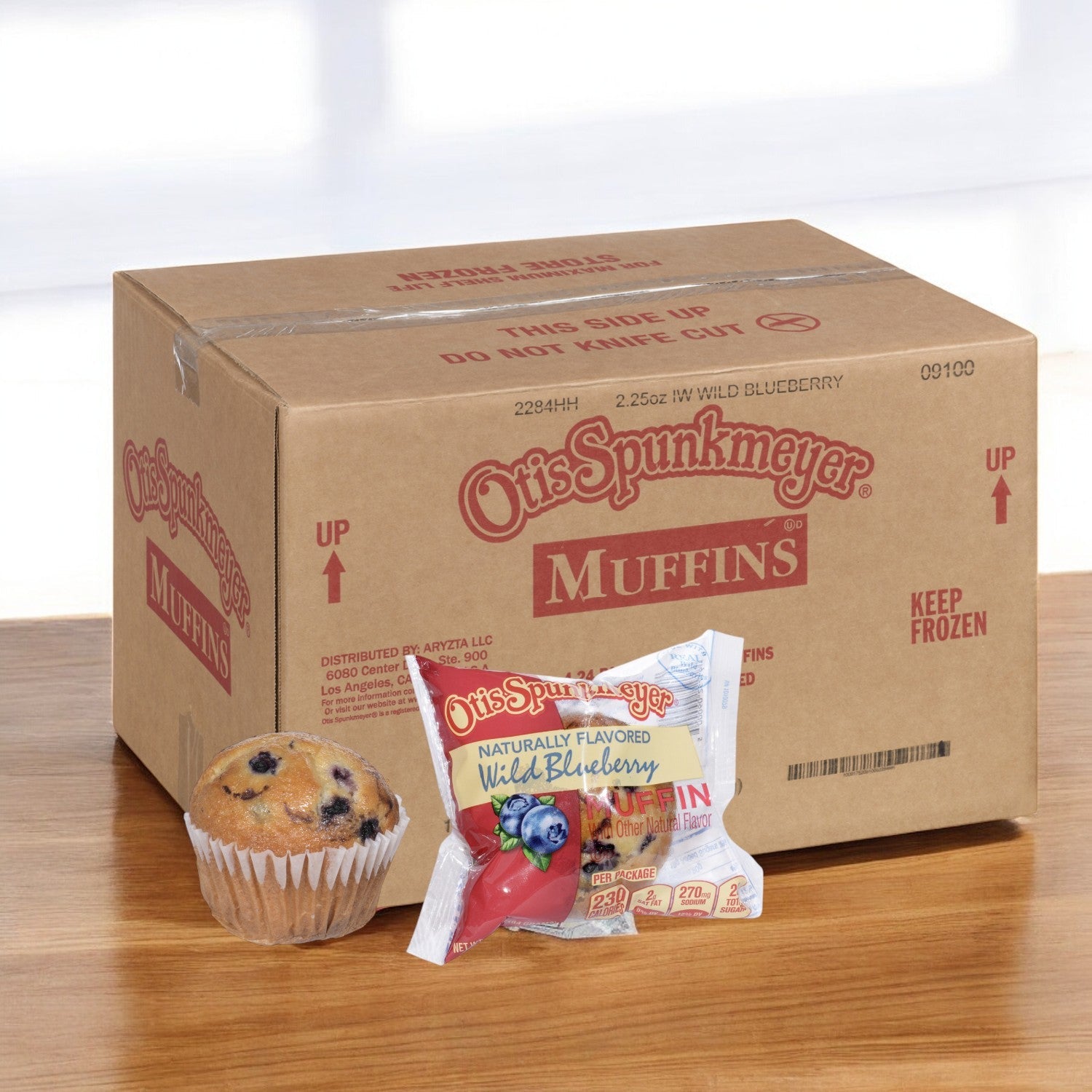A single Otis Spunkmeyer Muffin Blueberry, from a case of 96 individually wrapped frozen muffins, is displayed in its package on a wooden surface.