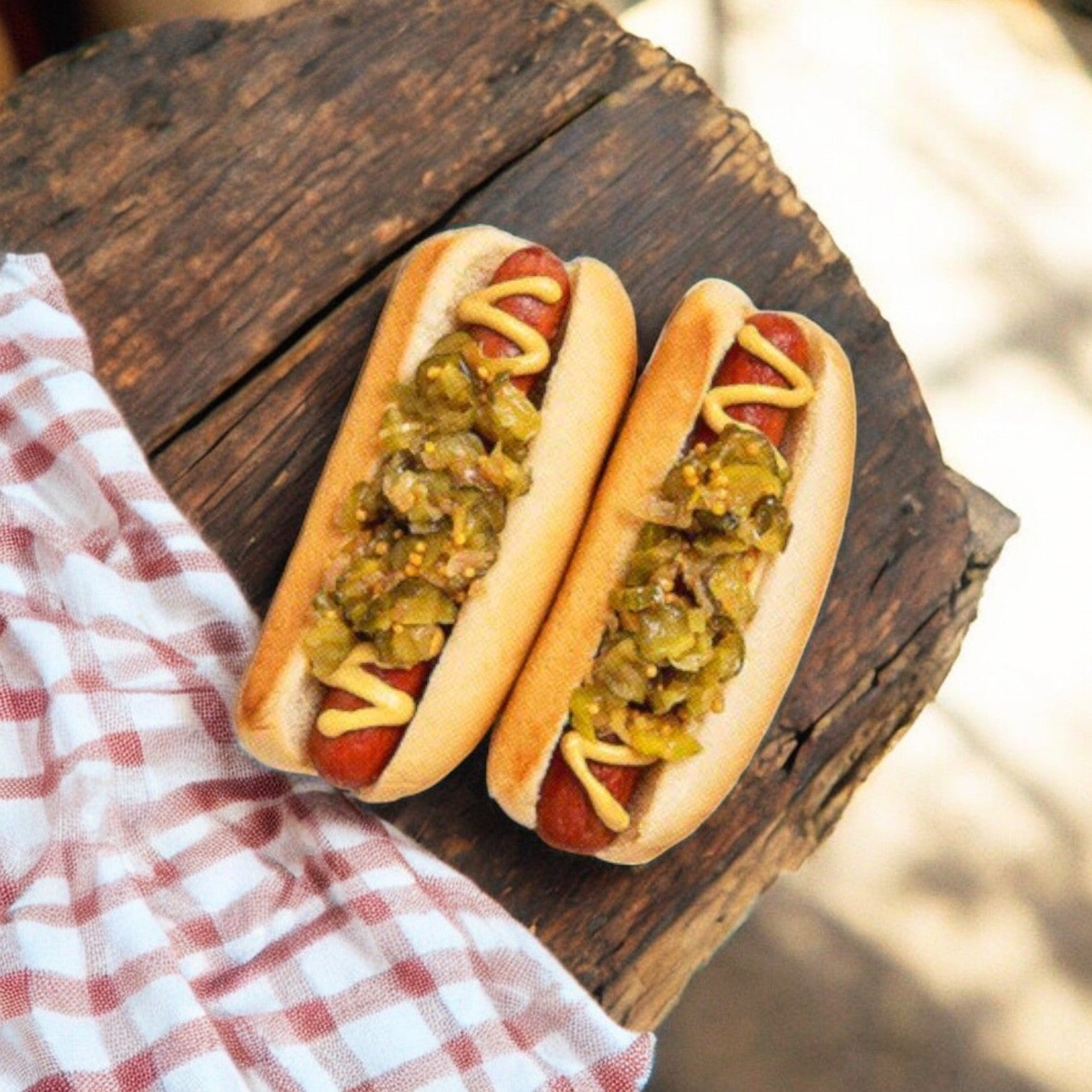Two Nathan’s Famous Angus Beef Franks are topped with mustard and relish, situated on a wooden surface beside a red and white checkered cloth.