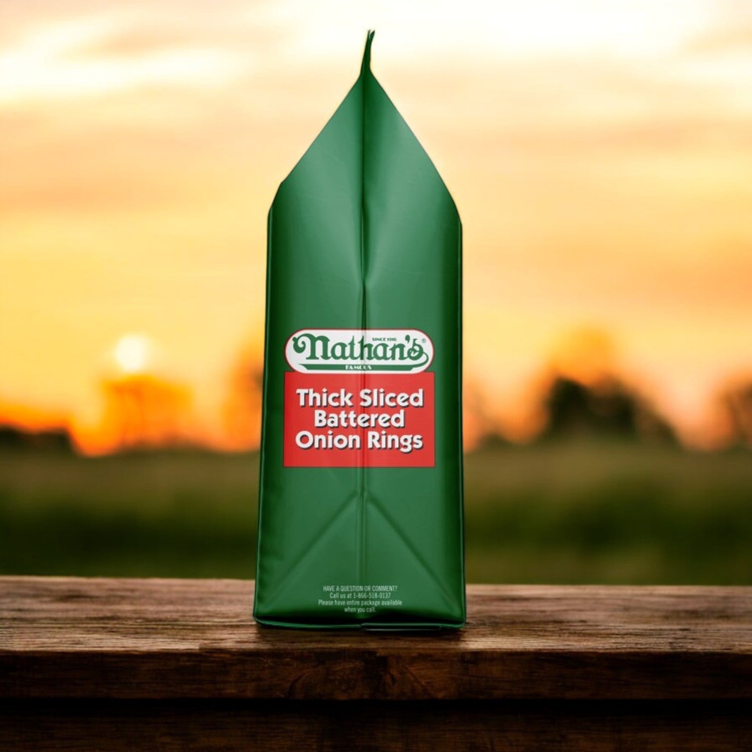 A package of Nathan’s Famous Onion Rings, Battered, Thick Sliced – 16 oz - 3 Count sits on a wooden surface, its green packaging highlighted against a sunset sky.