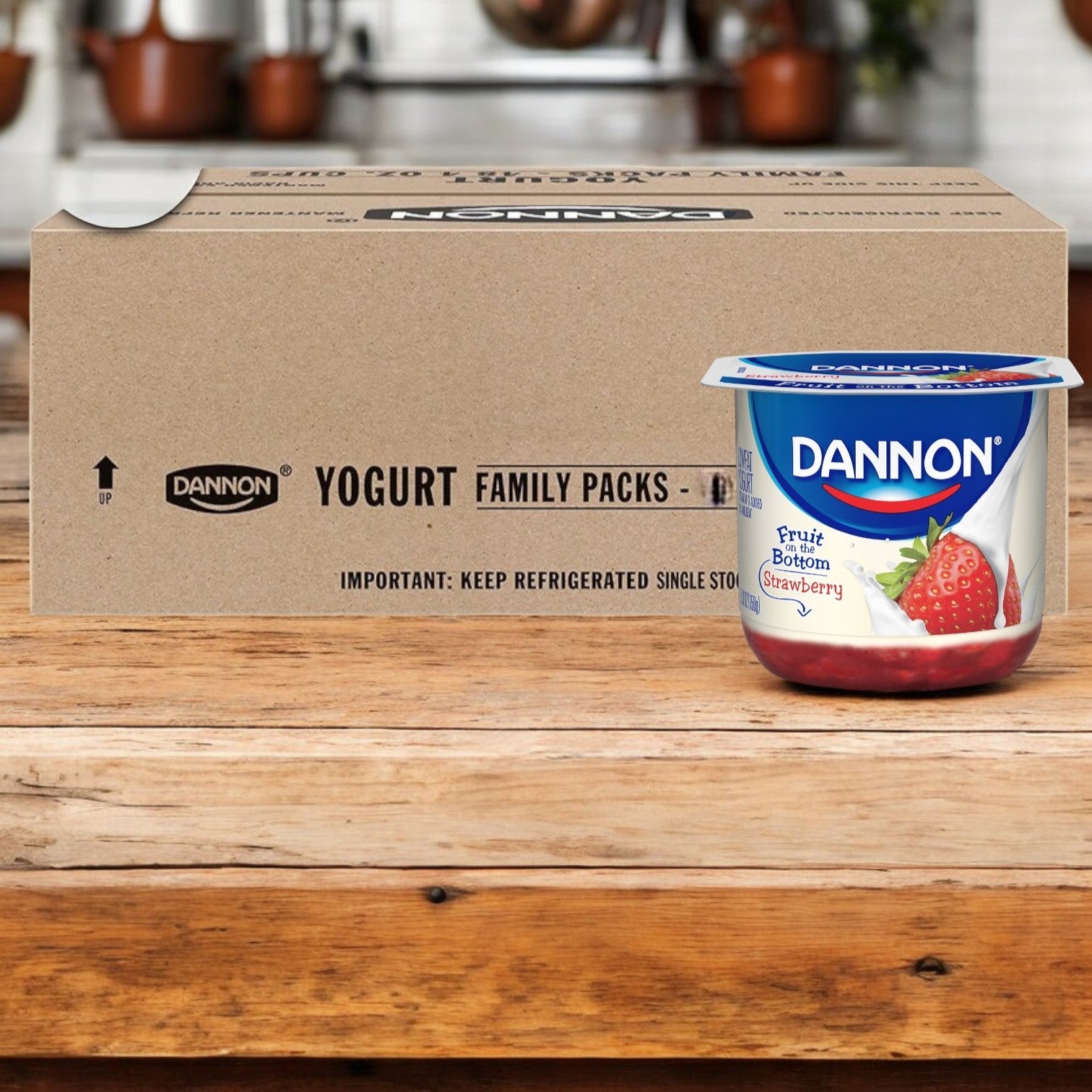 A container of Dannon Fruit on the Bottom Strawberry Low Fat Yogurt (5.3 oz.) is placed on a wooden surface in front of a case box of the same product, which contains 12 yogurts per case.