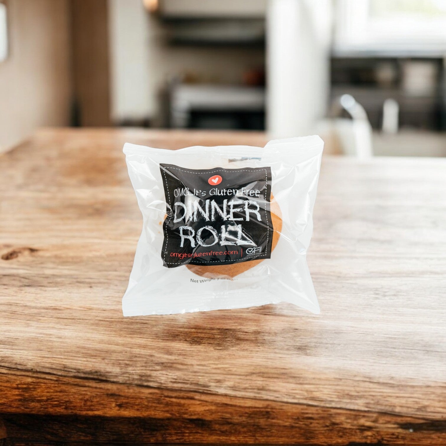 An OMG It's Gluten Free, Dinner Roll, 2 oz. - 1 Count, in clear packaging with a black label sits on a wooden table against a blurry kitchen background. The soft and fluffy texture promises deliciousness in every bite.
