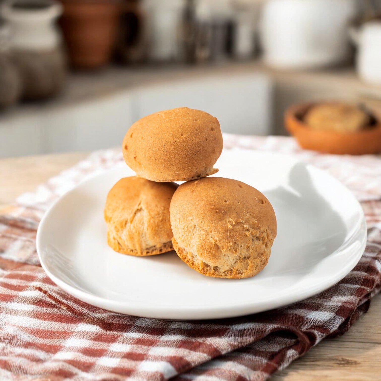 A single 2 oz. dinner roll from OMG It's Gluten Free is stacked on a white plate, placed on a red and white checkered cloth. The background shows a blurred kitchen setting.