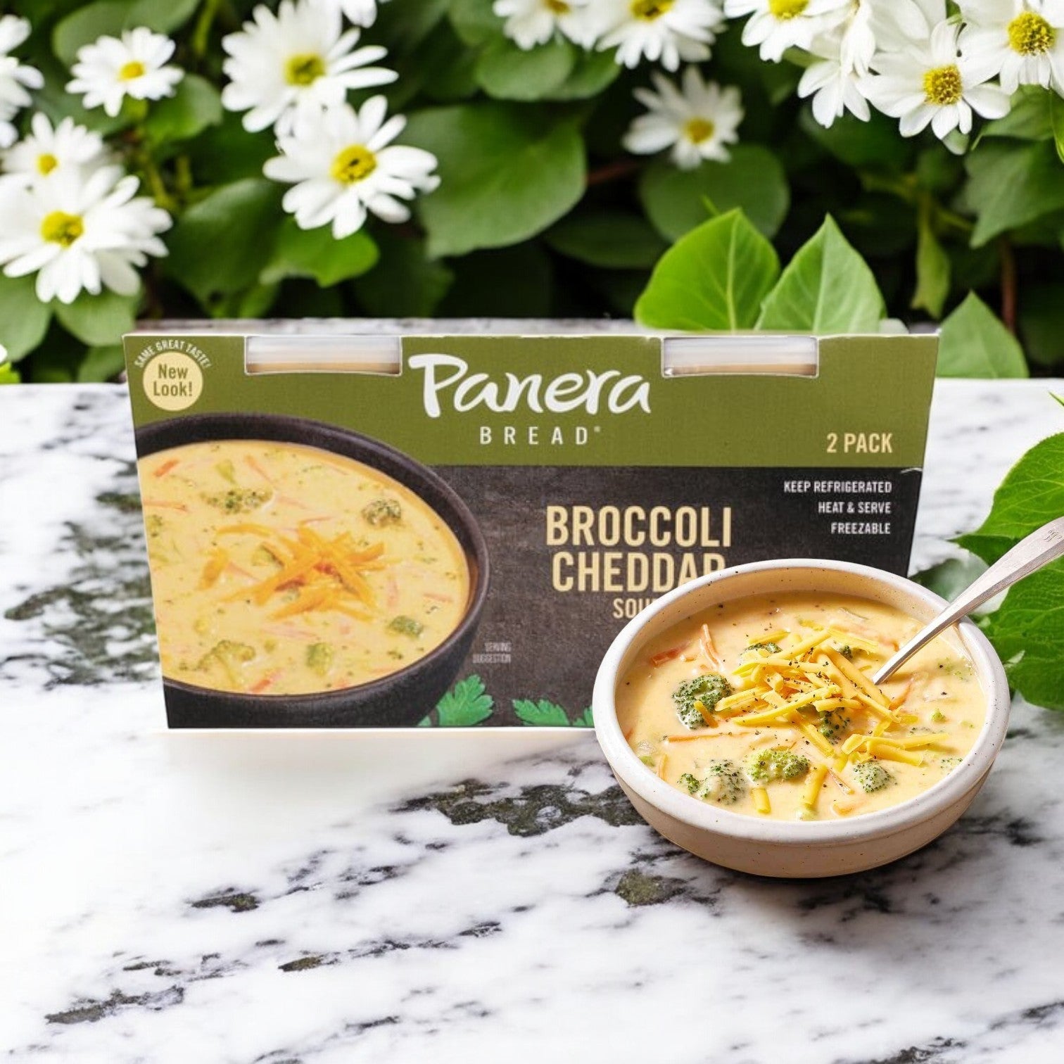 A bowl of creamy broccoli cheddar soup with a spoon is beside a Panera Bread Broccoli Cheddar Soup 24 oz, 2-Count package on marble, with white flowers and green leaves in the background.