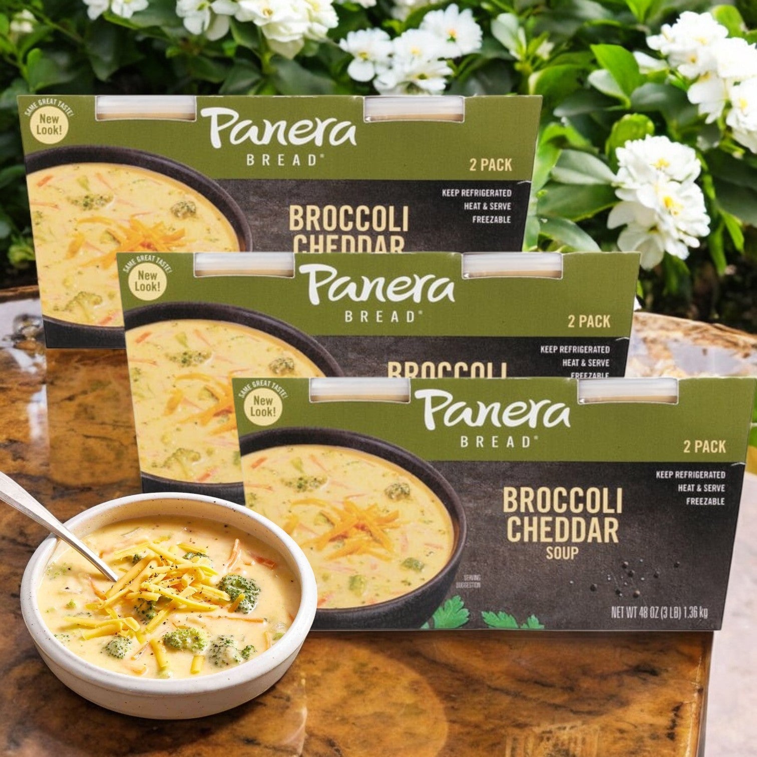 Three packs of Panera Bread’s Broccoli Cheddar Soup (each containing two 24 oz portions) are displayed outdoors, with a bowl garnished with shredded cheese and broccoli in front, ready to heat and enjoy.