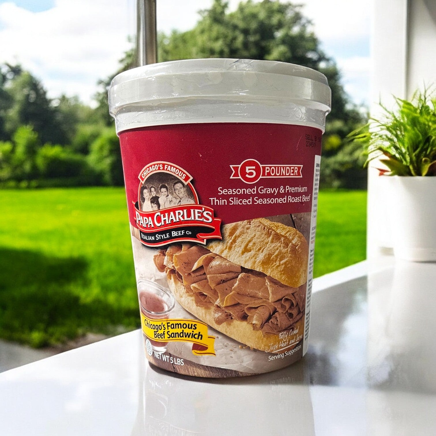 A Papa Charlie's 32 Sandwich Combo featuring seasoned roast beef with a sandwich image on the label rests on a white surface. Giardiniera peeks from behind hoagie rolls, while greenery and a tree are blurred in the background through a window. Perfect for parties with included buns!.