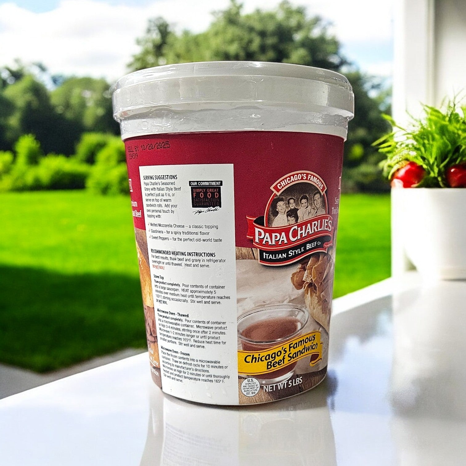 A container of Papa Charlie's Thin Sliced Roast Beef, seasoned and sliced, rests on a white table outside. In the background, a grassy lawn and potted plant create a peaceful atmosphere.
