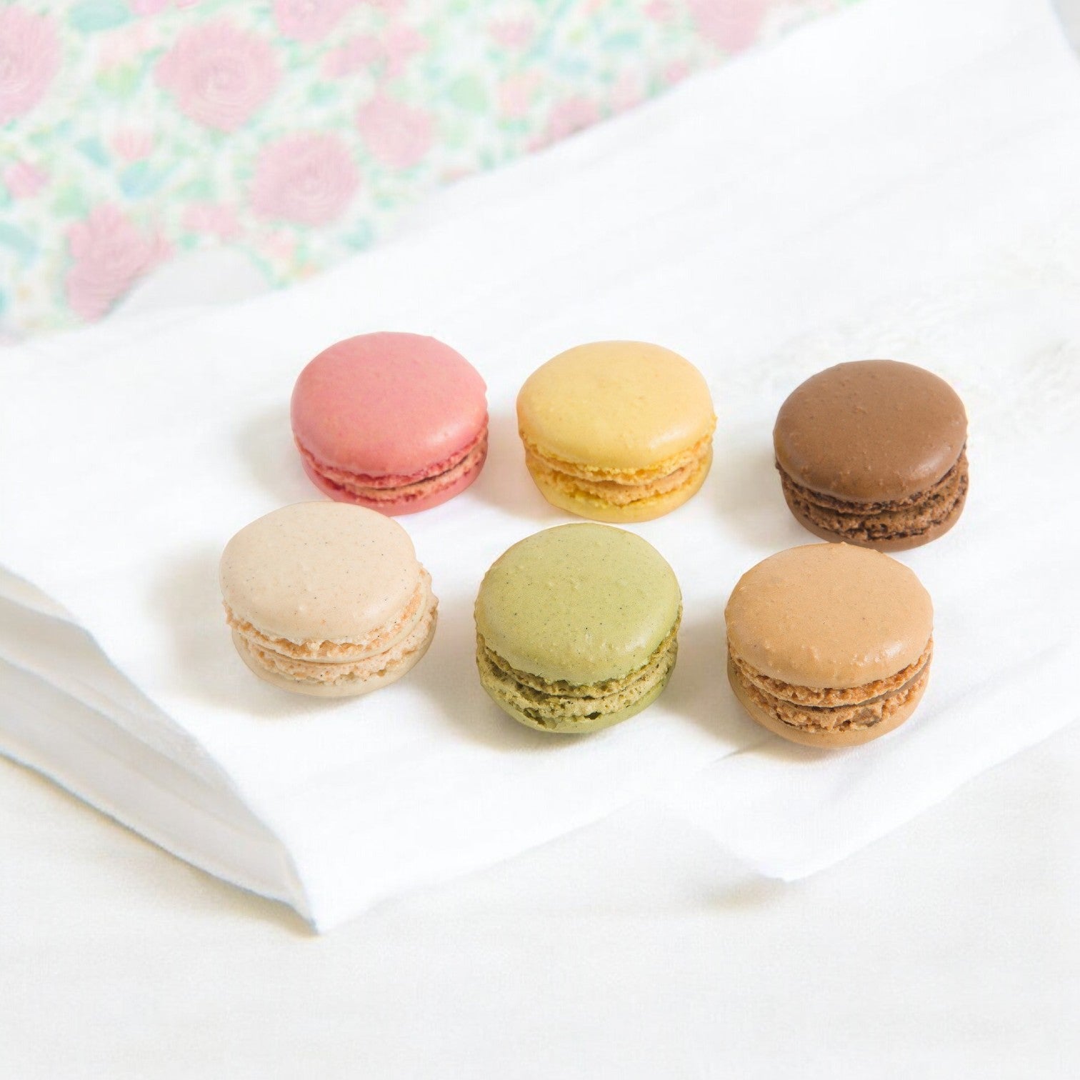 Pasquier Mini Assorted Macarons—featuring pink, yellow, brown, beige, and green hues—are elegantly displayed on white paper over a light surface. These treats offer authentic French craftsmanship in every bite. Enjoy 144 pieces with two trays of 72 each.