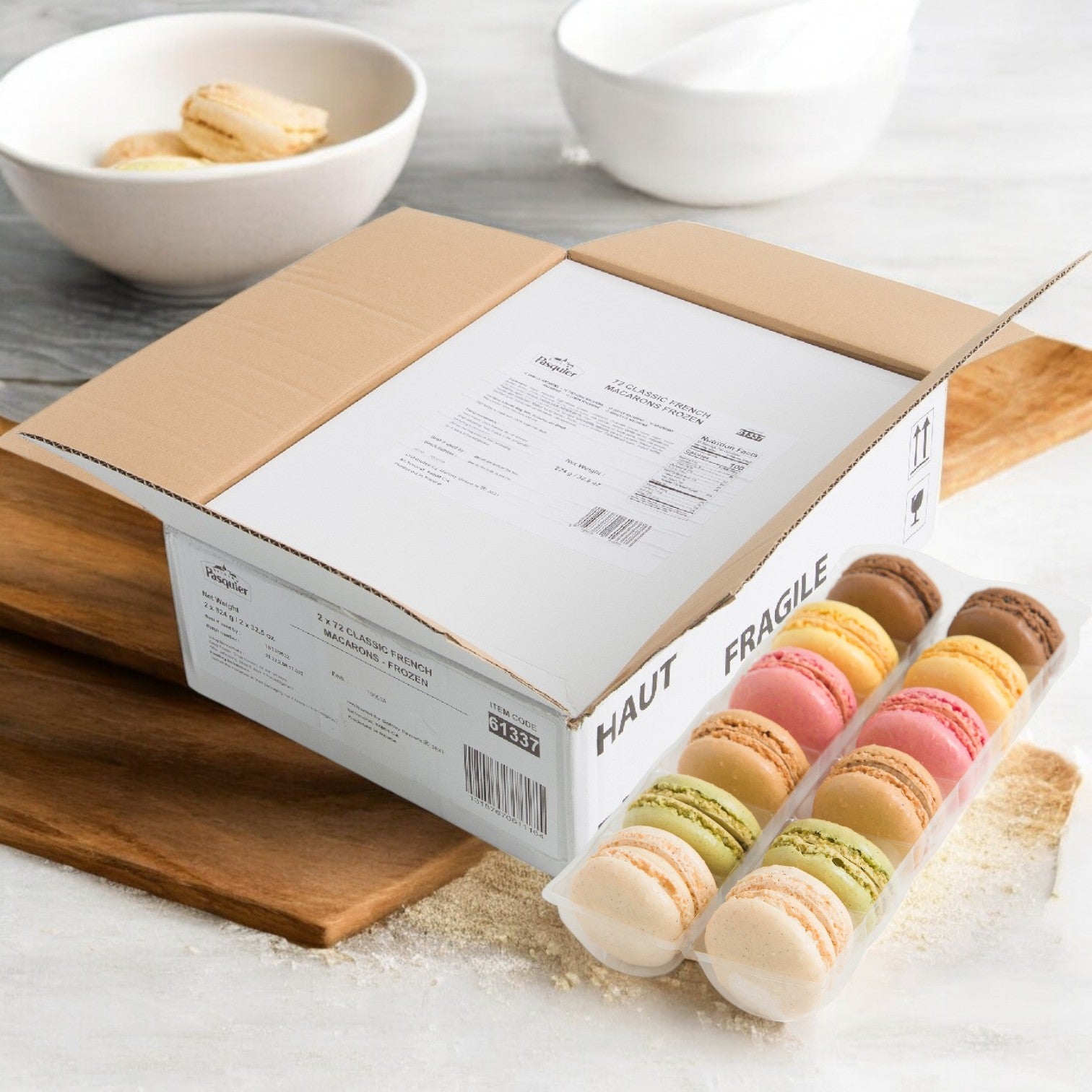 A "fragile" cardboard box sits on a wooden surface beside four rows of Pasquier Mini Assorted Macarons, 2 trays of 72 each, with a white bowl in the background, highlighting authentic French craftsmanship.