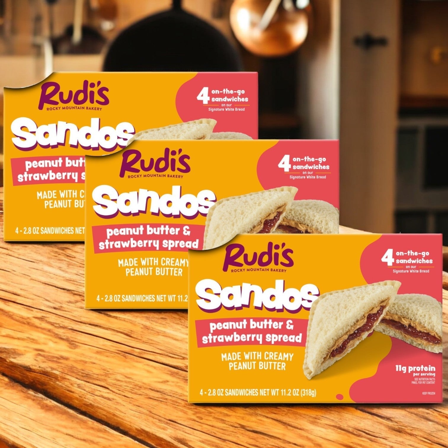 Rudi's Rocky Mountain Bakery's 3-pack Peanut Butter and Strawberry Spread Sandos, totaling 11.2 oz, are showcased on a wooden table. These vegan snacks offer four convenient, on-the-go sandwiches per box.