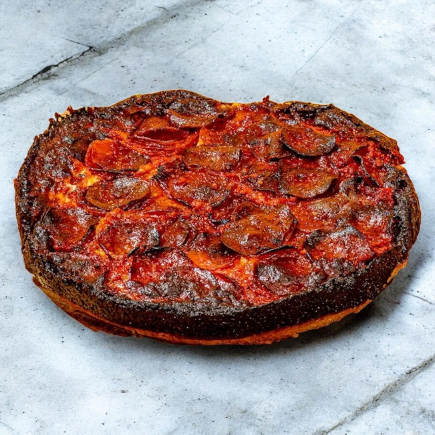 The rustic, round Pequods Pizza Pepperoni, 2" tall with a thick, charred crust topped with pepperoni and melted cheese enticingly sits on a gray stone surface, reminiscent of a delightful creation from Pequod Pizza.