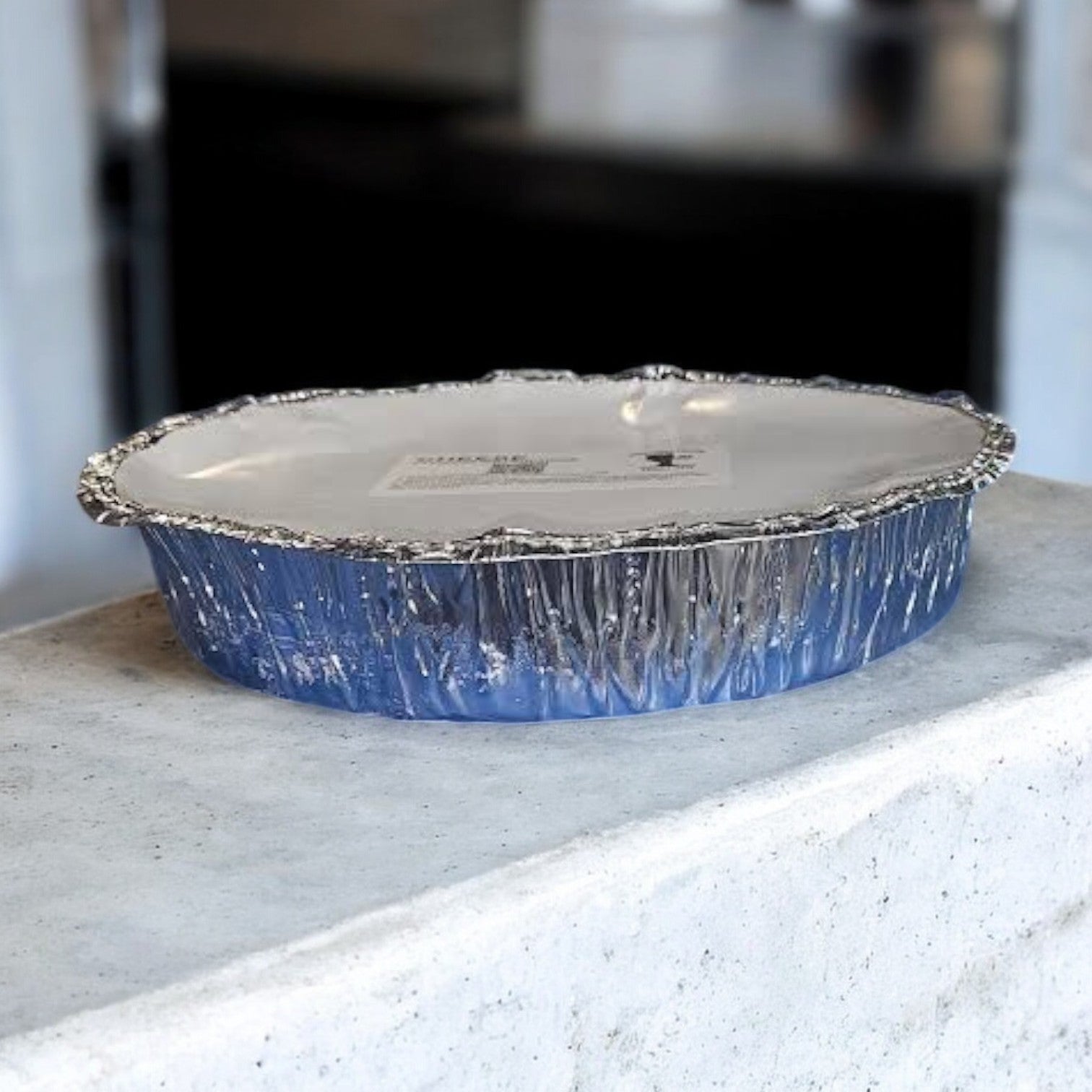 A round, foil baking dish, like Pequod Pizza's deep dish style, rests on a light surface with a matching lid.