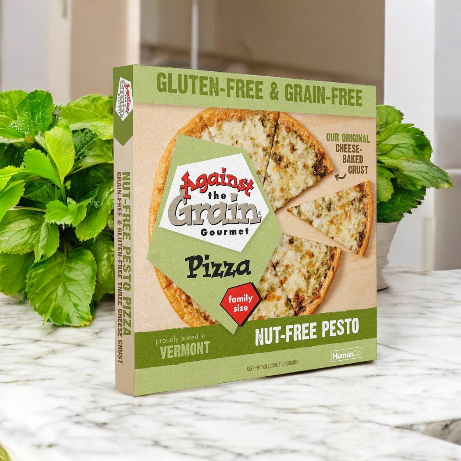 A box of Against the Grain Gourmet Pizza, Nut-Free Pesto, Family Size – 22.4 oz- 3 Packs rests on a marble counter with fresh greens. The packaging highlights its gluten-free crust and family-size serving for sharing.
