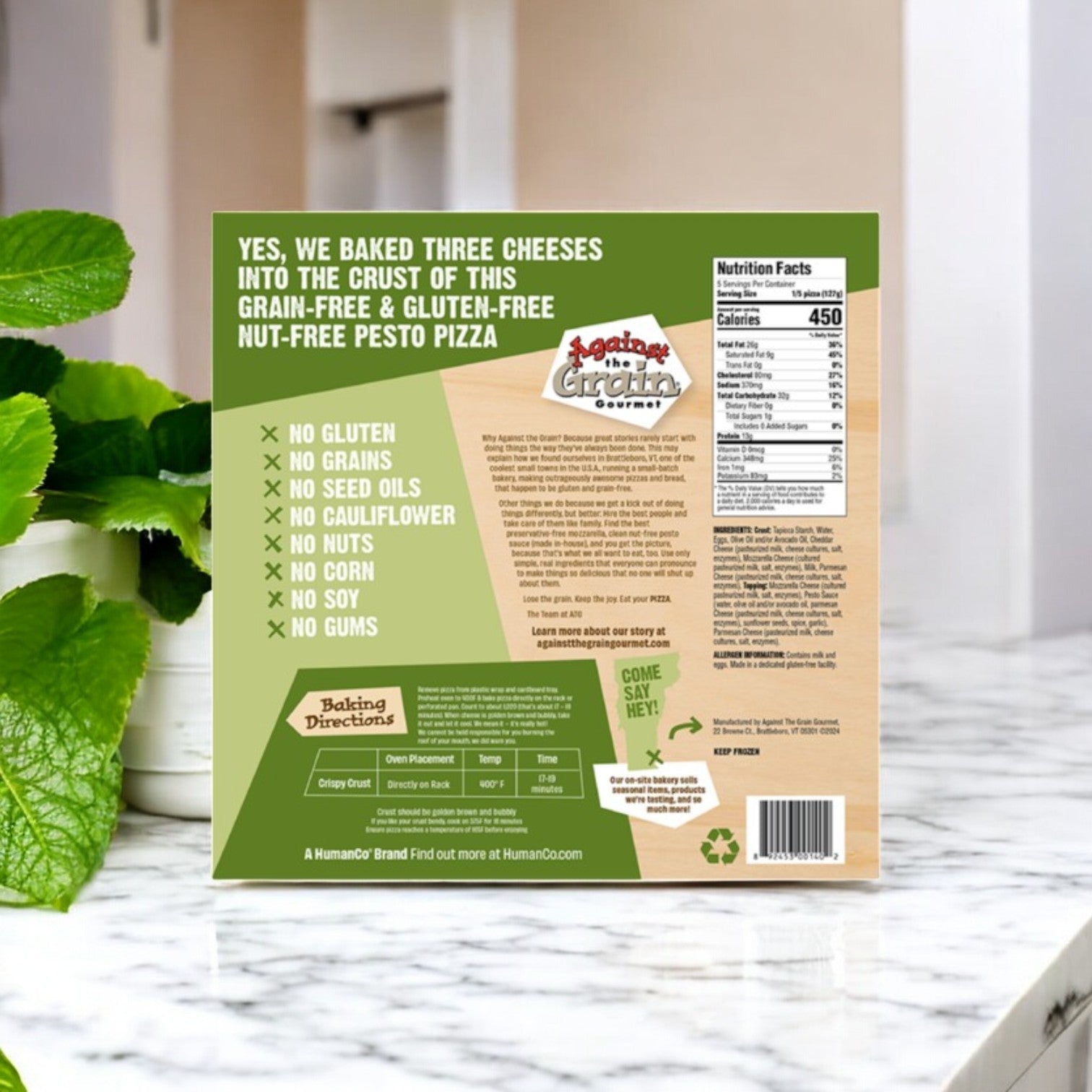 The Against the Grain Gourmet Pizza box, featuring a family-size nut-free pesto and gluten-free crust, is displayed elegantly on a marble countertop beside a vibrant green plant. Nutritional facts and baking directions are clearly printed on the 22.4 oz package.