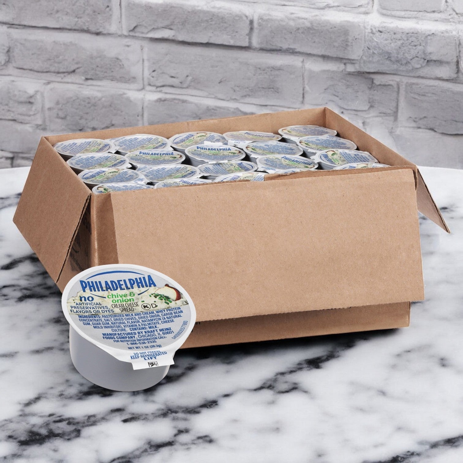 An open cardboard box displays Kraft's Philadelphia Chive & Onion Spread cups, elegantly placed on a marble surface with a brick wall backdrop. This 100-count pack offers endless culinary possibilities.