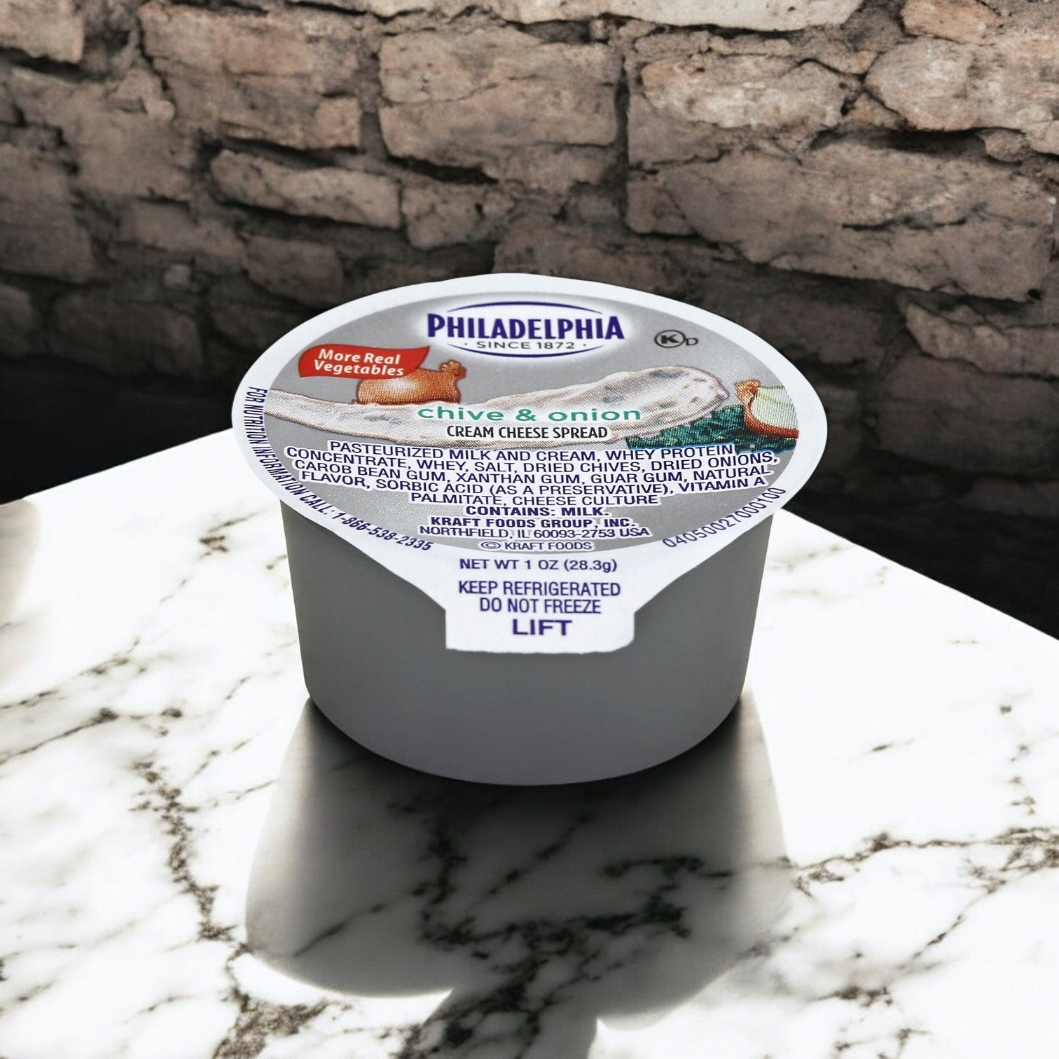 A 1 oz. cup of Kraft's Philadelphia Cheese Cream Chive & Onion Spread rests on a marble counter with a brick wall backdrop, highlighting its creamy richness.