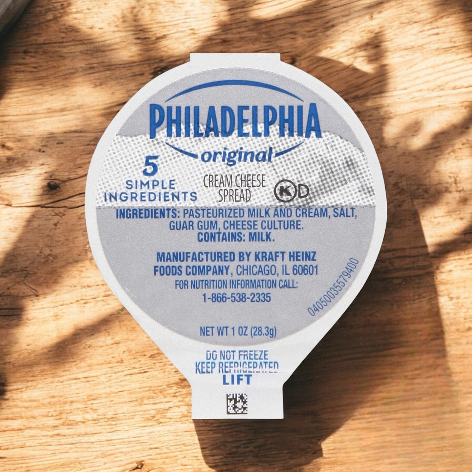 The Kraft Philadelphia Cream Cheese Original Spread, a 1 oz (28.3g) cup from a 100 count package, sits on a wooden surface displaying its ingredients, with "Do not freeze" clearly marked on the lid.