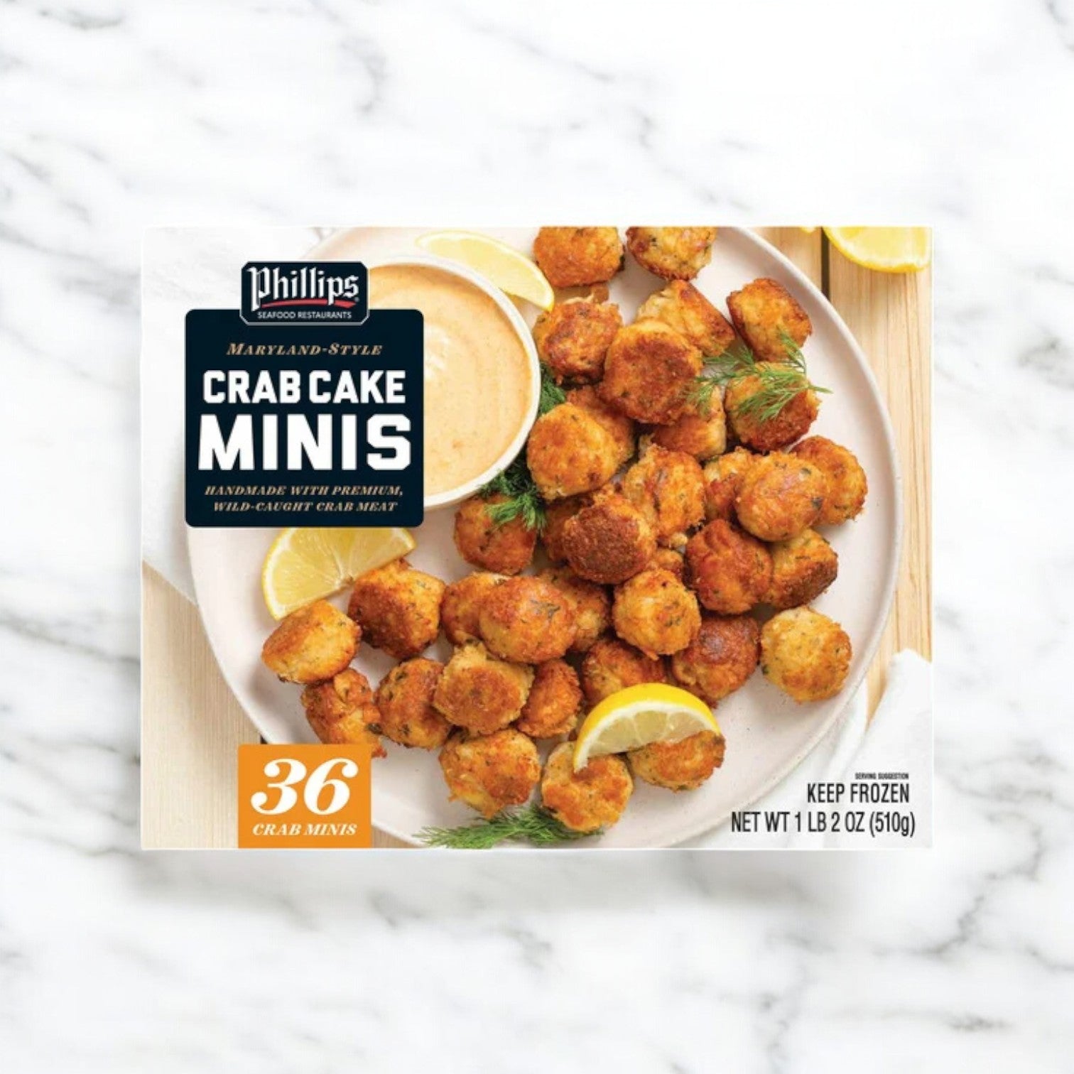 A box of Phillips Crab Cake Minis from Phillips Foods, featuring lemon wedges and dill, rests on a marble surface. It contains 36 mini crab cakes with a net weight of 1 lb 2 oz (510g).