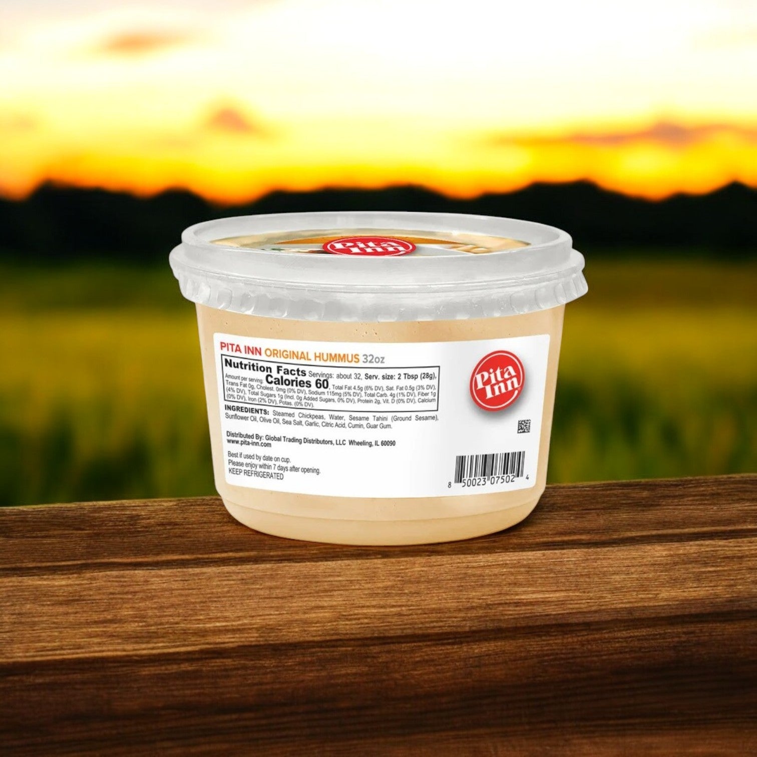 A 2 lb container of Good Foods' Pita Inn Original Hummus, boasting an authentic Mediterranean flavor, rests on a wooden surface against a blurred sunset backdrop.