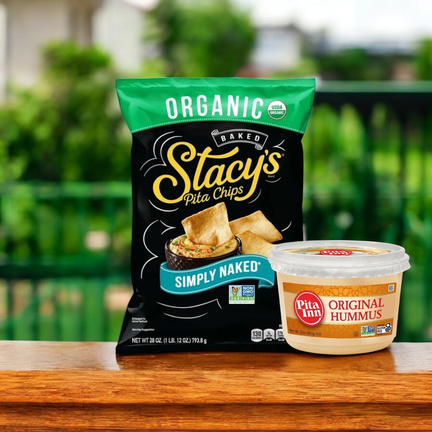 The Pita Inn Hummus & Stacy’s Pita Chips Combo sits elegantly on a wooden surface, offering a delightful Mediterranean taste against a blurred green backdrop.