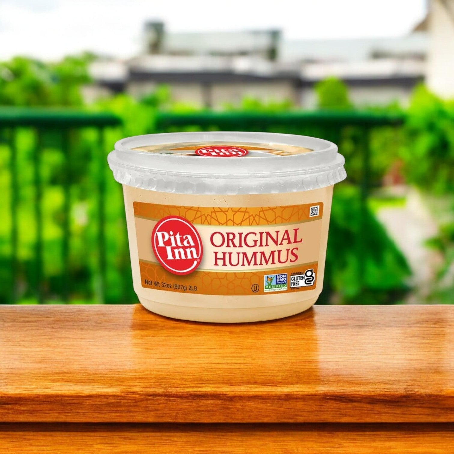 A container of Pita Inn Hummus & Stacy’s Pita Chips Combo – The Perfect Pair!! sits on wood, surrounded by blurred greenery and a fence, embodying that rich Mediterranean taste in an inviting outdoor setting.