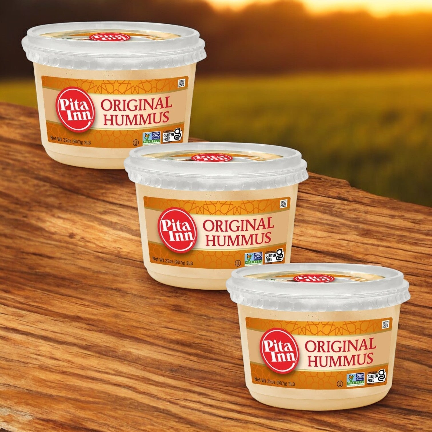 Three tubs of Good Foods' Pita Inn Original Hummus – Authentic Mediterranean Flavor, 2 lbs., are placed on a wooden surface, against a blurred outdoor backdrop.