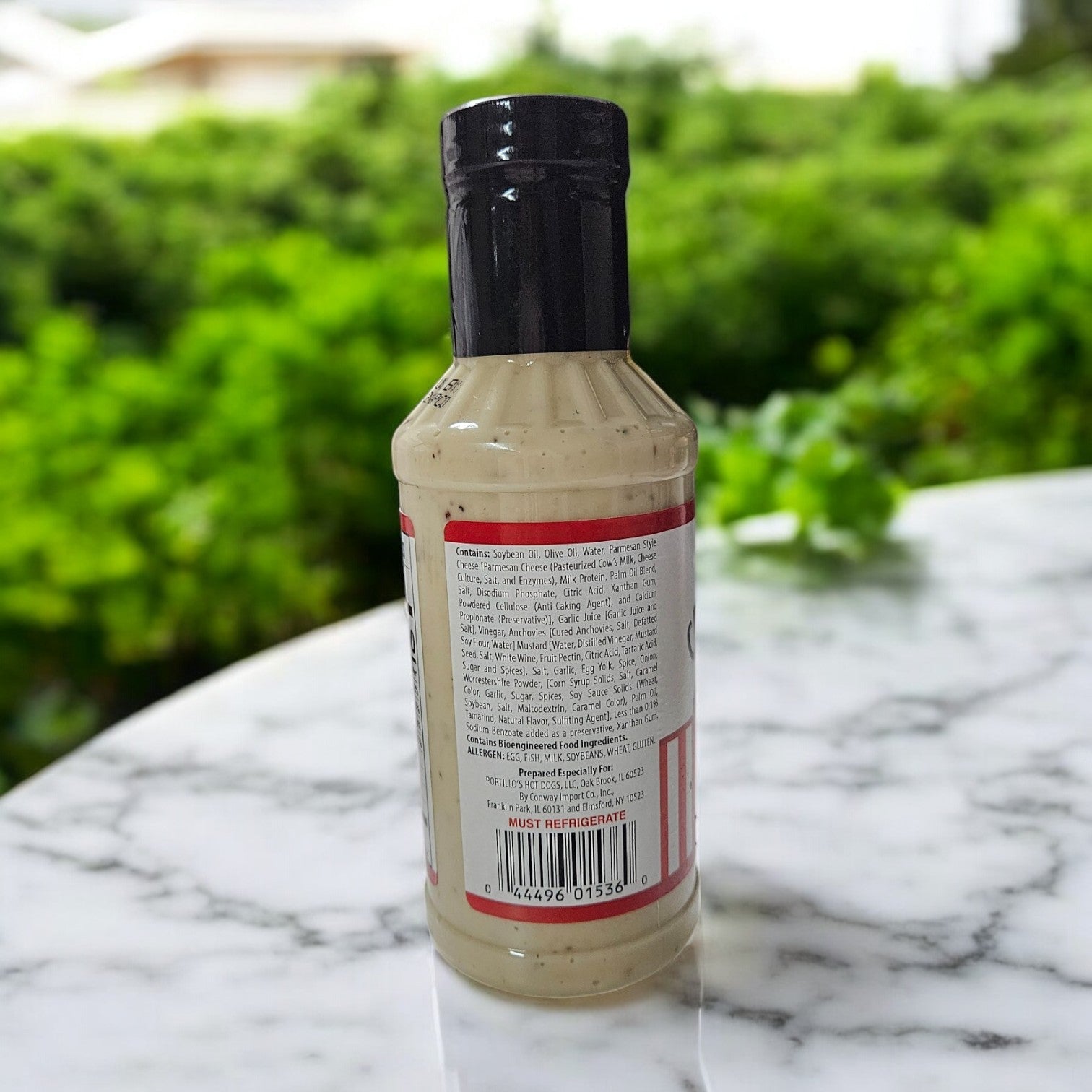 A 16 oz. Portillo's Caesar Dressing bottle sits on a marble table with its ingredients showing, against a backdrop of blurred greenery and buildings, evoking the freshness of a crisp Caesar salad.