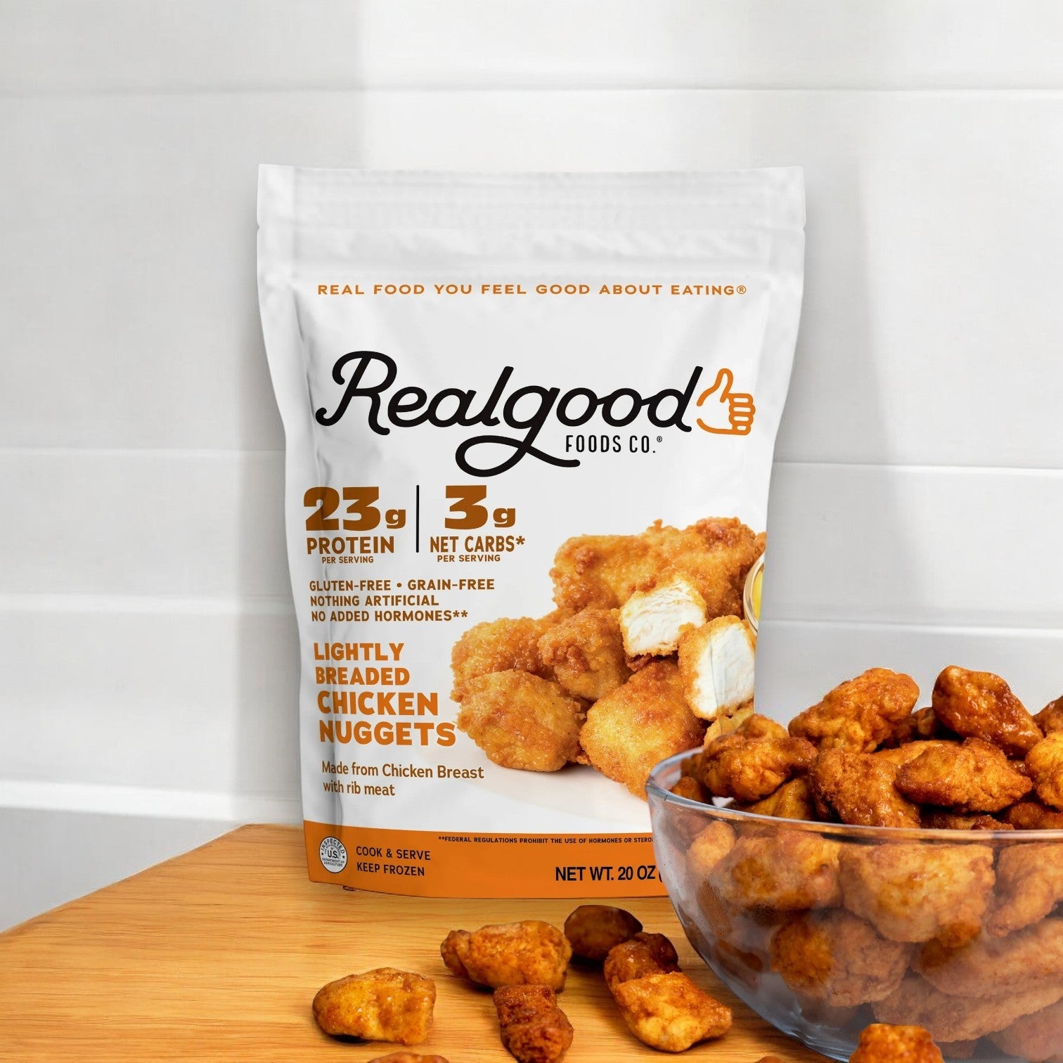 A 20 oz bag of Realgood Foods Co. Lightly Breaded Chicken Breast Nuggets sits on the countertop, with a few crispy nuggets resting in a glass bowl nearby.