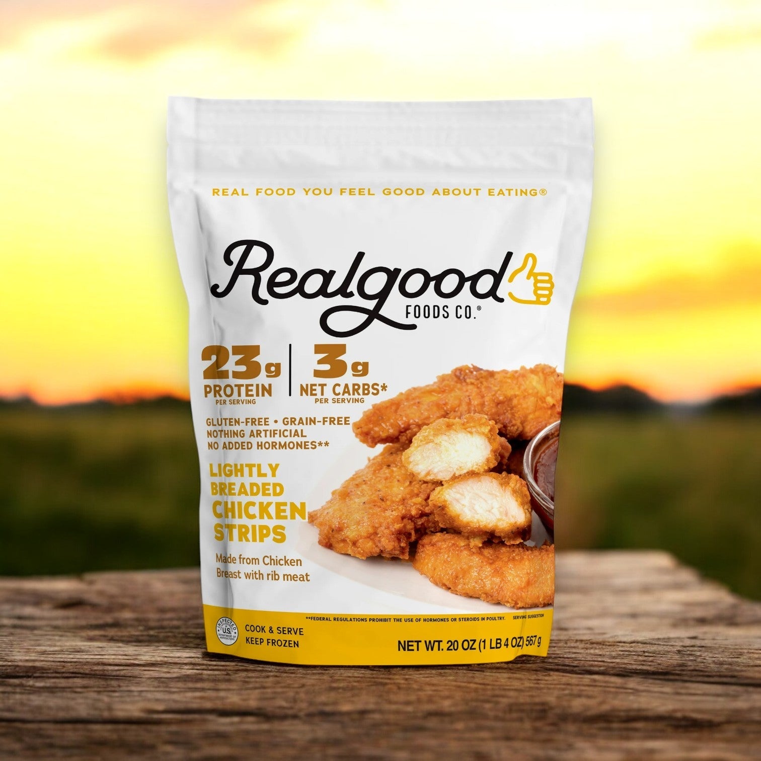 A 20 oz bag of Real Good Foods Lightly Breaded Chicken Breast Strips, gluten-free, rests on a wooden surface with a blurred outdoor background.