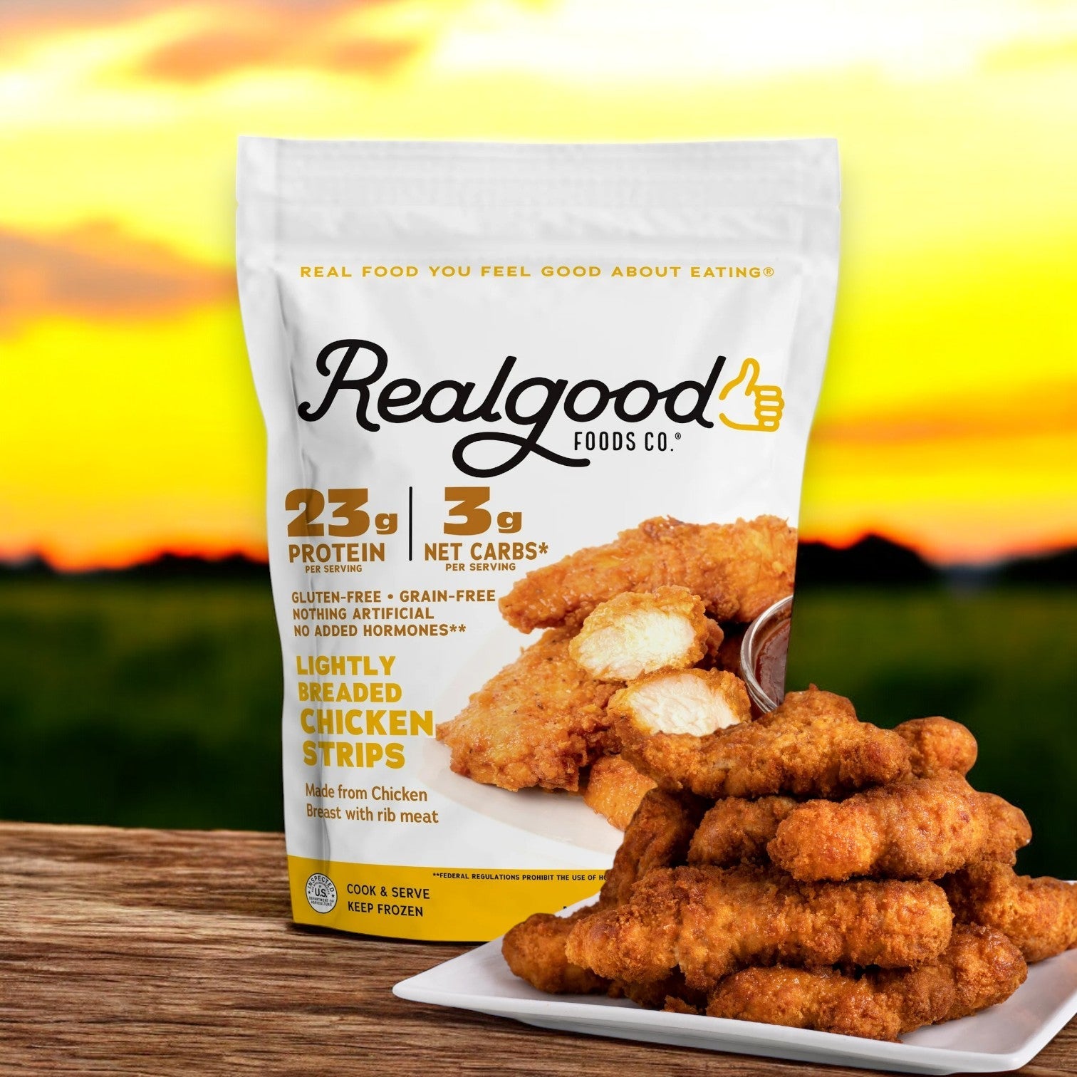 A package of Real Good Foods Lightly Breaded Chicken Breast Strips, 20 oz, is shown with a plate of the gluten-free strips in front, set against a sunset backdrop. The packaging emphasizes premium chicken breast and nutritional benefits.