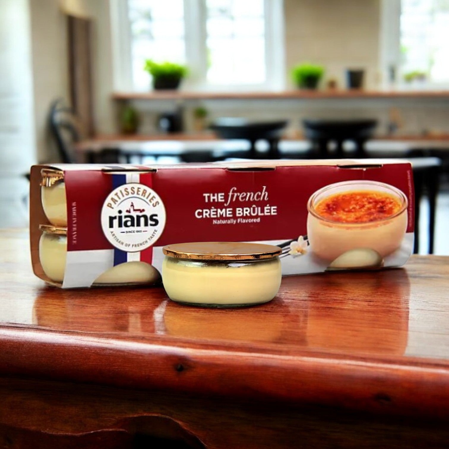 A package of Rian's La Crème Brûlée, 18 pk – Decadent French Dessert, is showcased on a wooden table with a single jar of vanilla custard. A blurred kitchen serves as the backdrop.