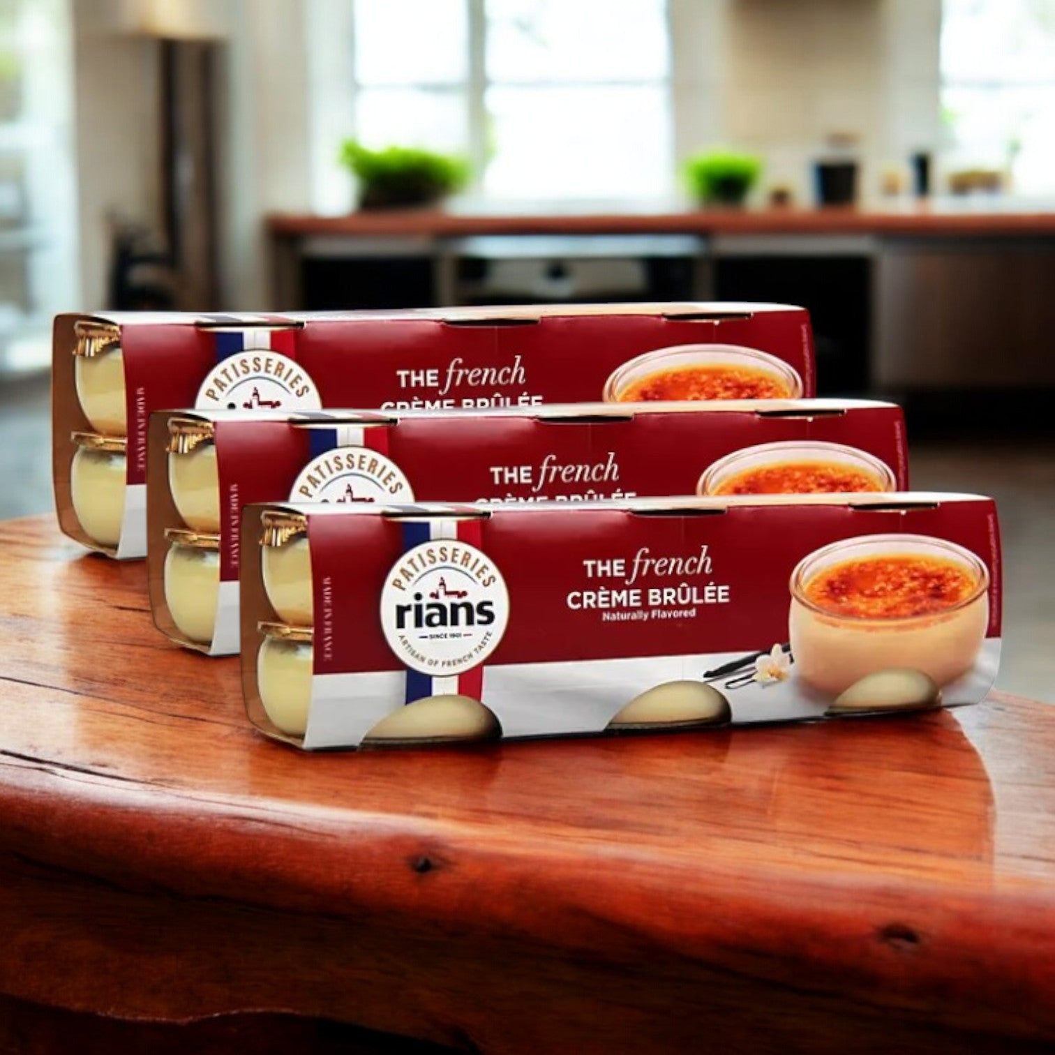 Three packs of Rian's La Crème Brûlée, an indulgent French vanilla custard dessert, sit on a wooden surface with a blurred kitchen backdrop.