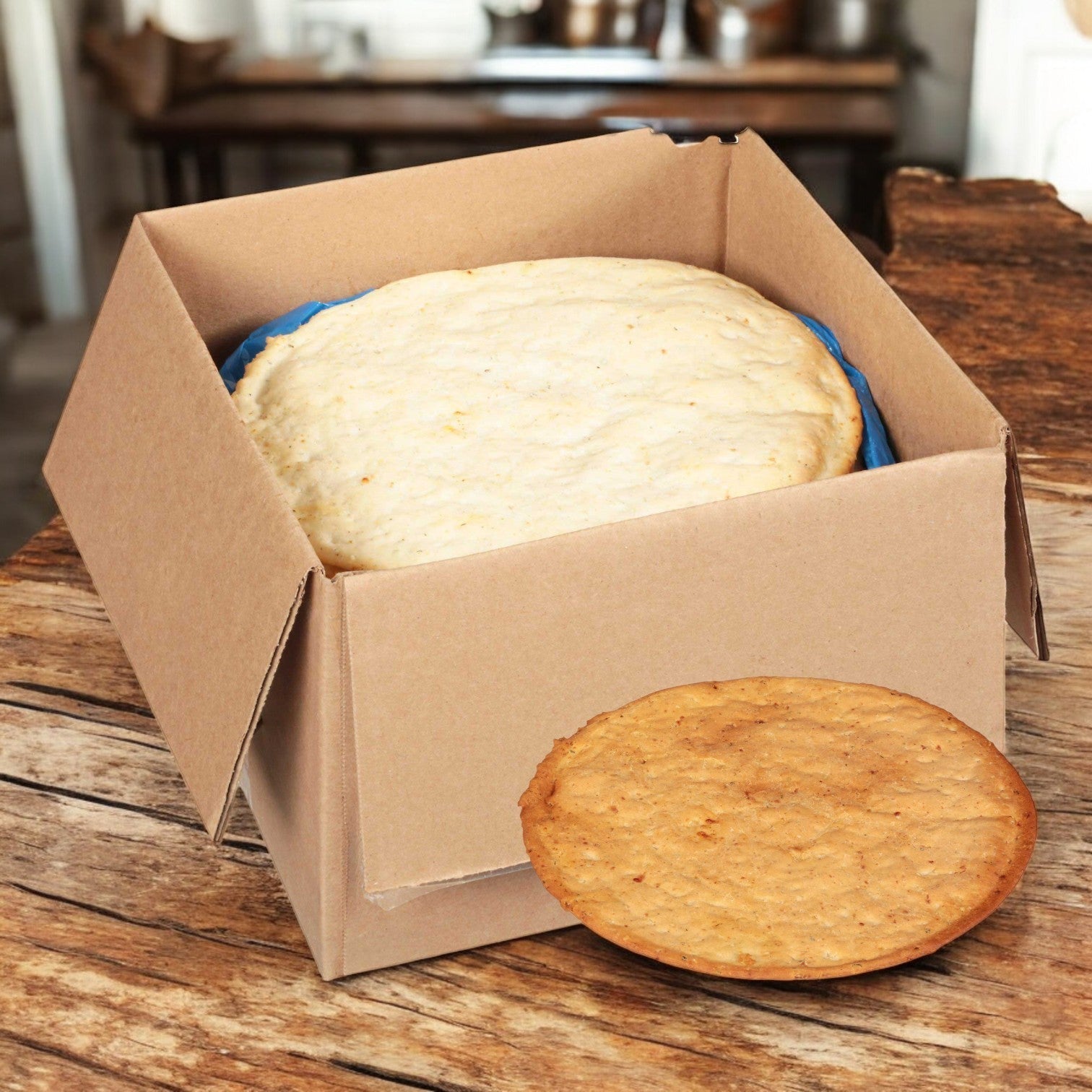 A large Rich's Gluten Free Seasoned Cauliflower Pizza Crust in an open cardboard box sits on a wooden table, with a smaller gluten-free biscuit in front of the box.