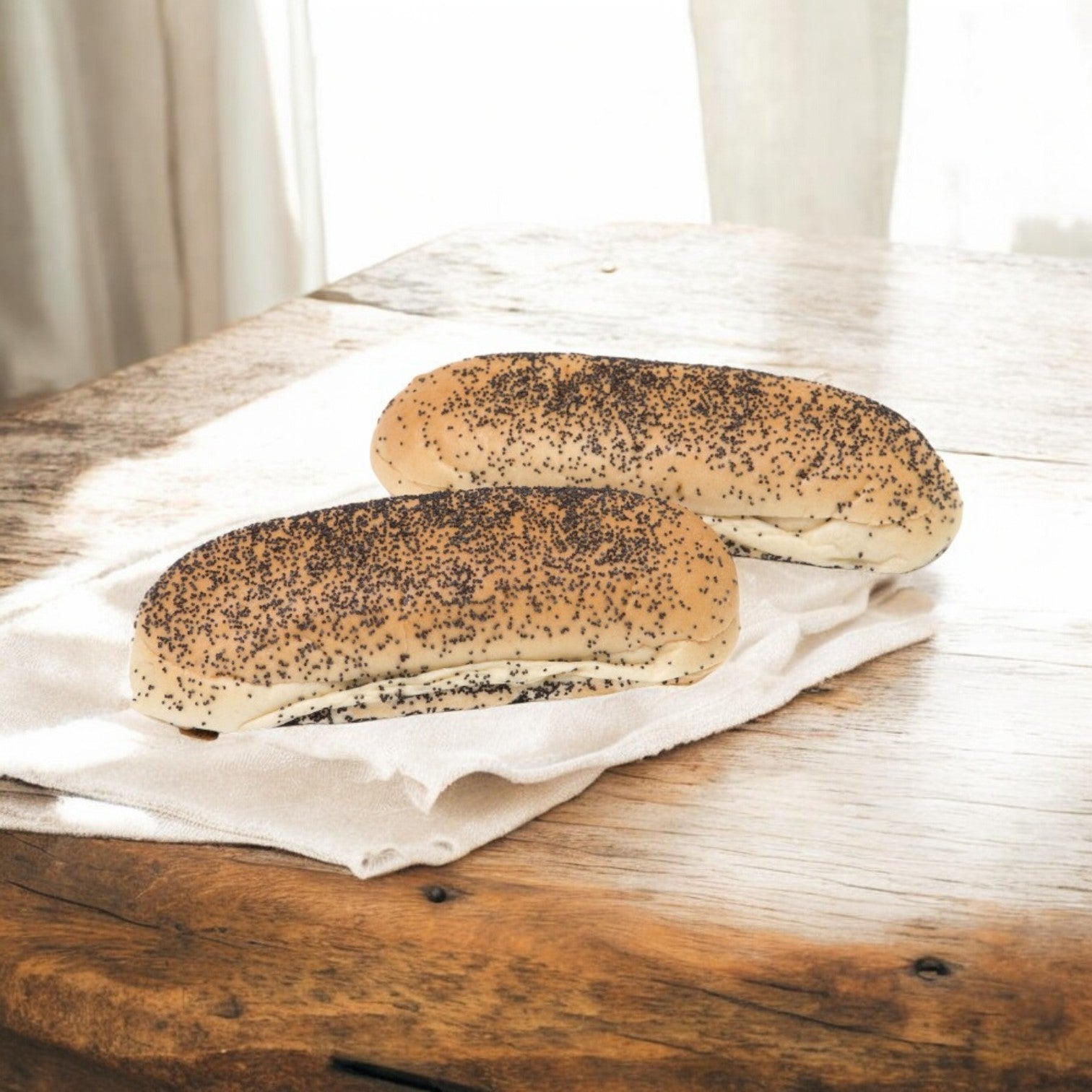 Two Rotellas Poppyseed Sliced Hot Dog Buns from Easy Lunches rest on a white cloth on a sunlit wooden table. These 5.5" artisanal delights, reminiscent of soft deli buns, are ready to elevate any meal.