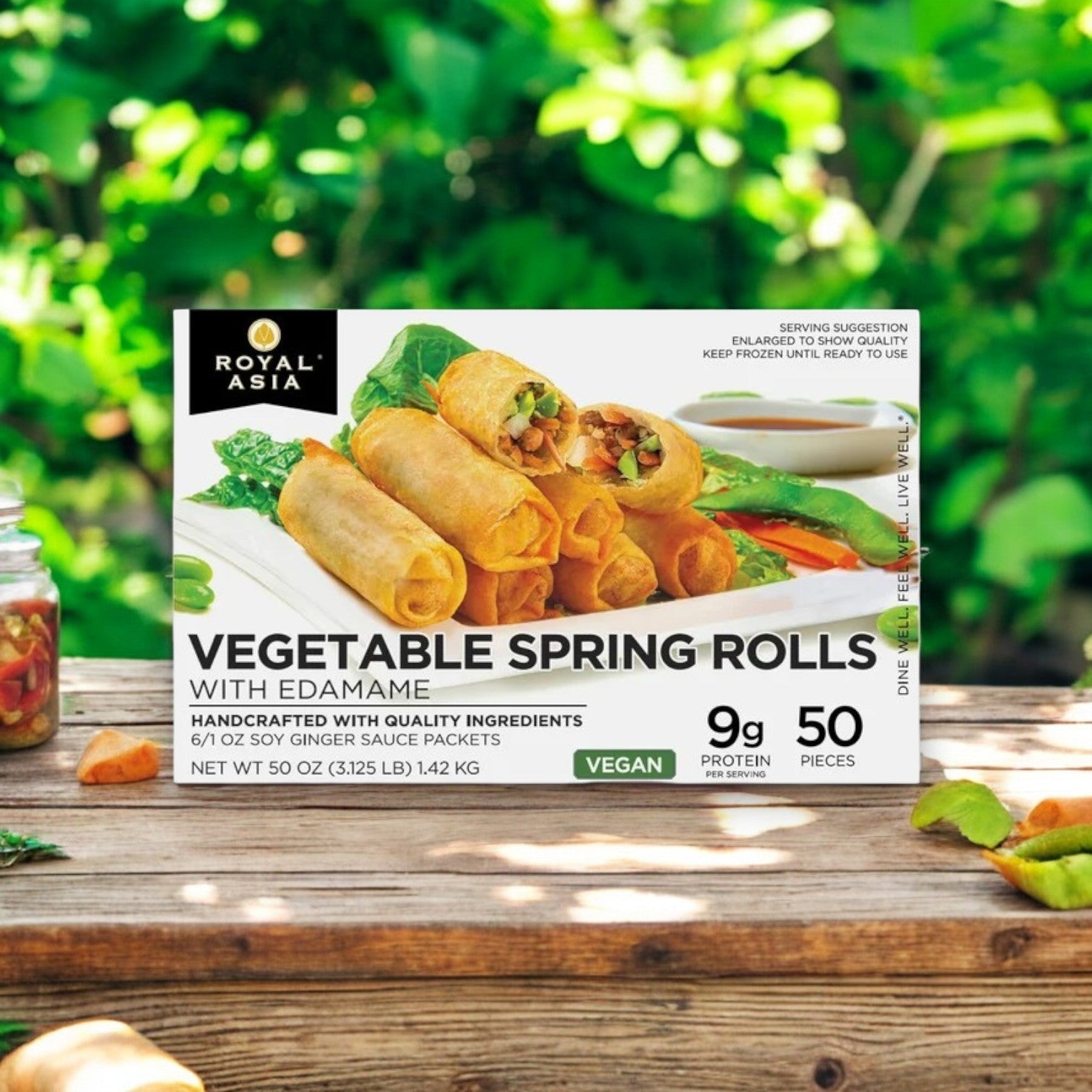 A 50-count box of Bibigo's Royal Asia Vegetable Spring Rolls with Soy Ginger Sauce, containing 9g protein and weighing 48 oz, sits on a wooden table surrounded by greenery.