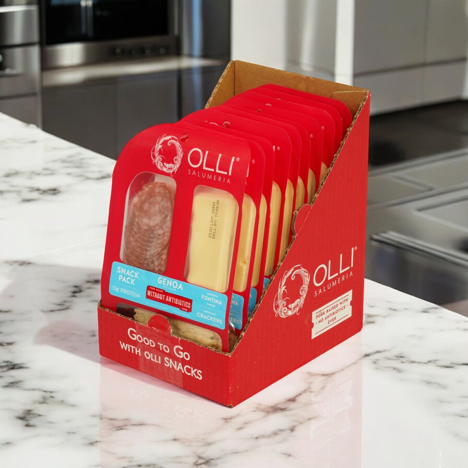 A red Olli display box elegantly sits on the marble countertop, showcasing Salami Genoa Fontina Snack Packs. These offer a tantalizing taste of Italy with their trio of Genoa salami, fontina cheese, and crackers.