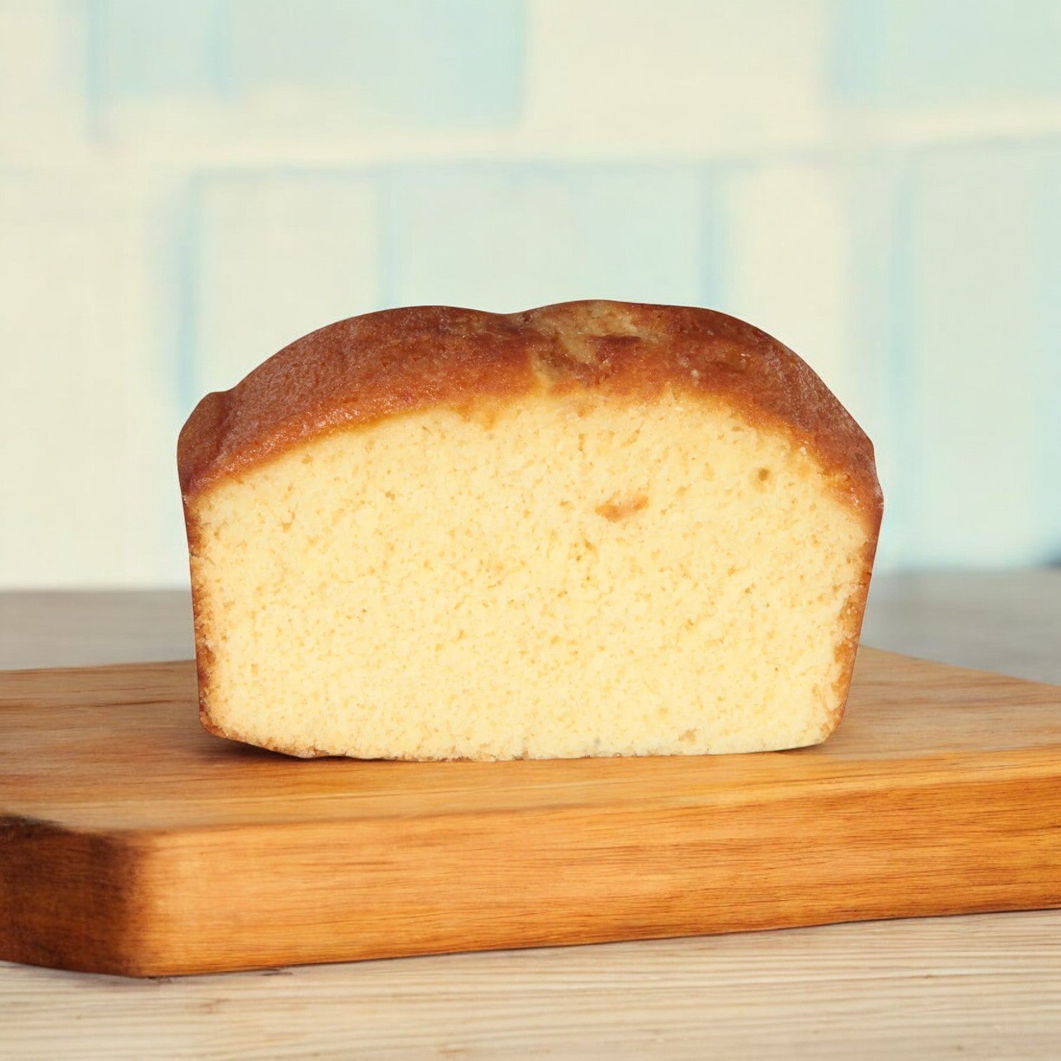 A Sara Lee Pound Cake, known for its moist texture and buttery flavor, is elegantly sliced on a wooden cutting board. Its light brown crust and pale interior contrast the blurred blue background.