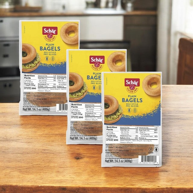 Three Schar Bagels, Gluten-Free, New York Style, Plain, Pre-Sliced packages are on a wooden table. The yellow packaging details product and nutritional info, ideal for those with gluten sensitivities craving authentic New York bagels.