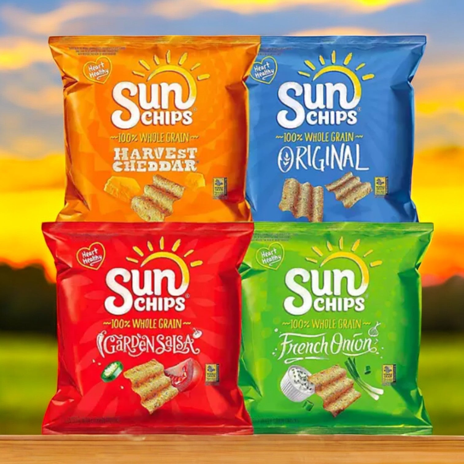 Four SunChips bags from the SunChips Variety Pack Whole Grain Chips, 30 pk., are displayed against a sunset backdrop: Harvest Cheddar (orange), Original (blue), Garden Salsa (red), and French Onion (green). Enjoy your favorite Sun Chips whole grain flavors.