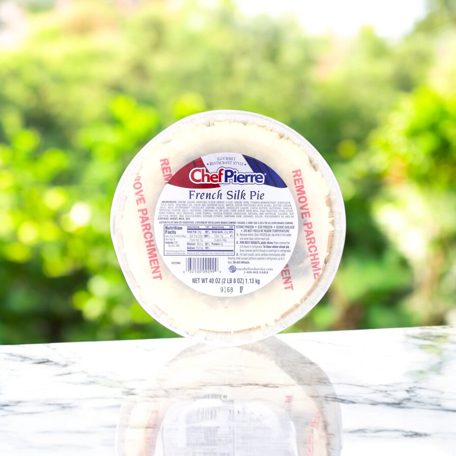 A round package of Chef Pierre's Chocolate French Silk Pie, 10-inch and 34 oz, sits on a marble surface with a blurred green background. This gourmet frozen dessert features labeled packaging and a warning to remove parchment before serving, highlighting its rich chocolate filling.