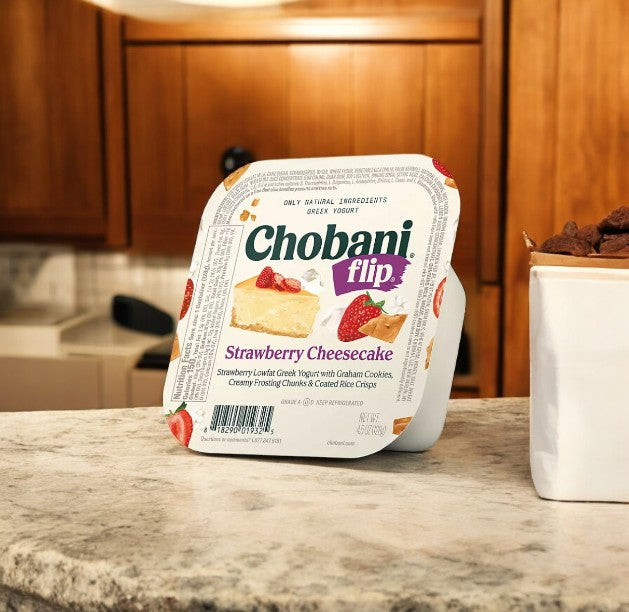 A Chobani Flip Lowfat Greek Yogurt from the variety pack rests on a marble countertop, with a cozy kitchen as the backdrop.