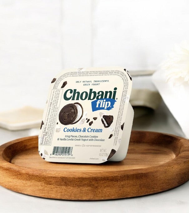 A Chobani Flip Lowfat Greek Yogurt container from the variety pack sits elegantly on a round wooden tray, set against a pristine white background.