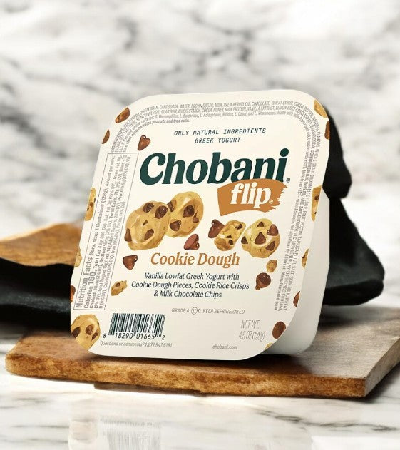 A Chobani Flip Lowfat Greek Yogurt Variety Pack, featuring cookie dough flavor, is placed on a marble surface with a wooden board. The packaging displays enticing cookie dough and chocolate chip images, ideal for savoring in this delightful 16 count, 4.5 oz pack from Chobani.