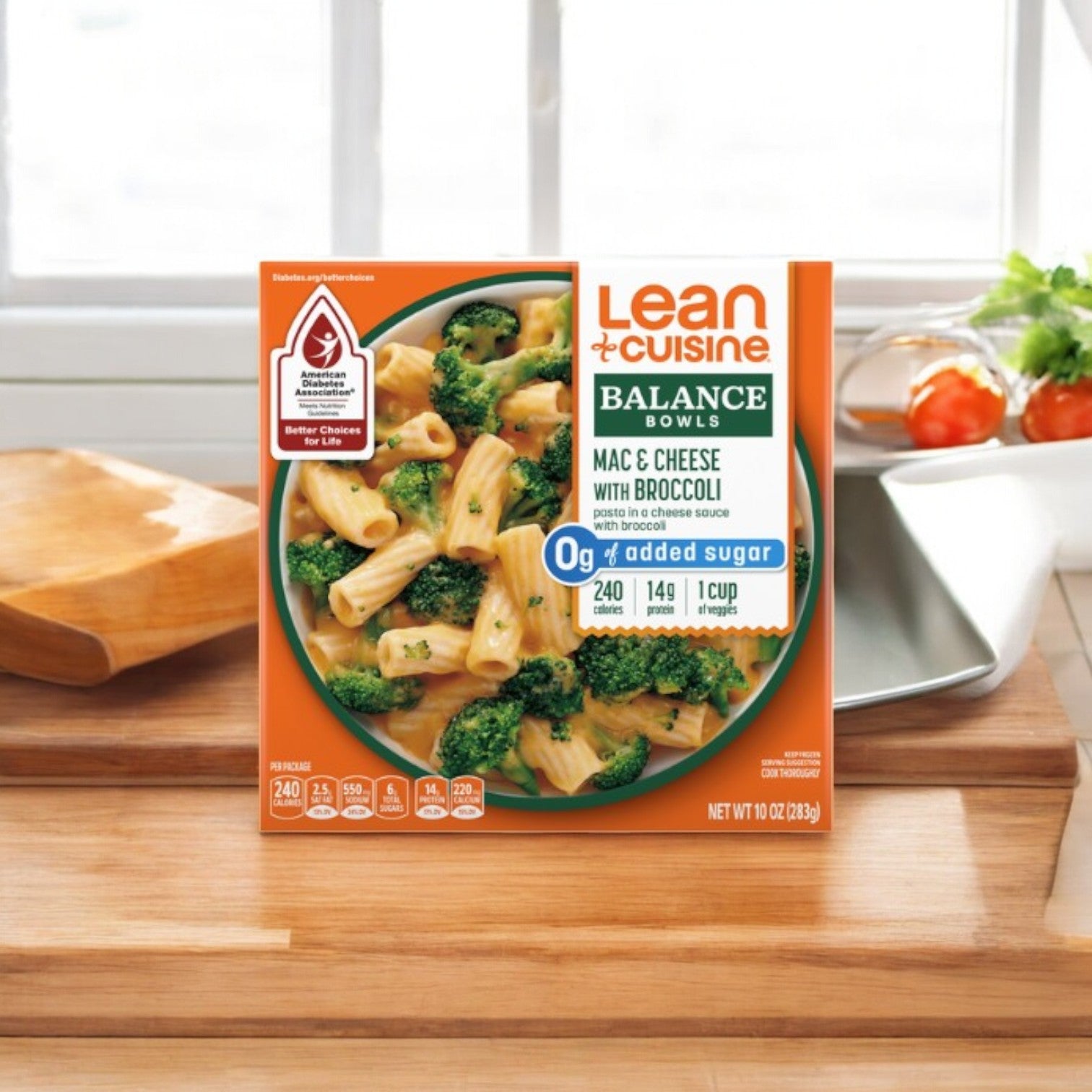 On a wooden counter sits a Lean Cuisine Mac & Cheese Broccoli Bowl, 10 oz. This dish includes creamy mac & cheese with vibrant broccoli. The packaging highlights nutritional information and proudly states there is 0g added sugar.