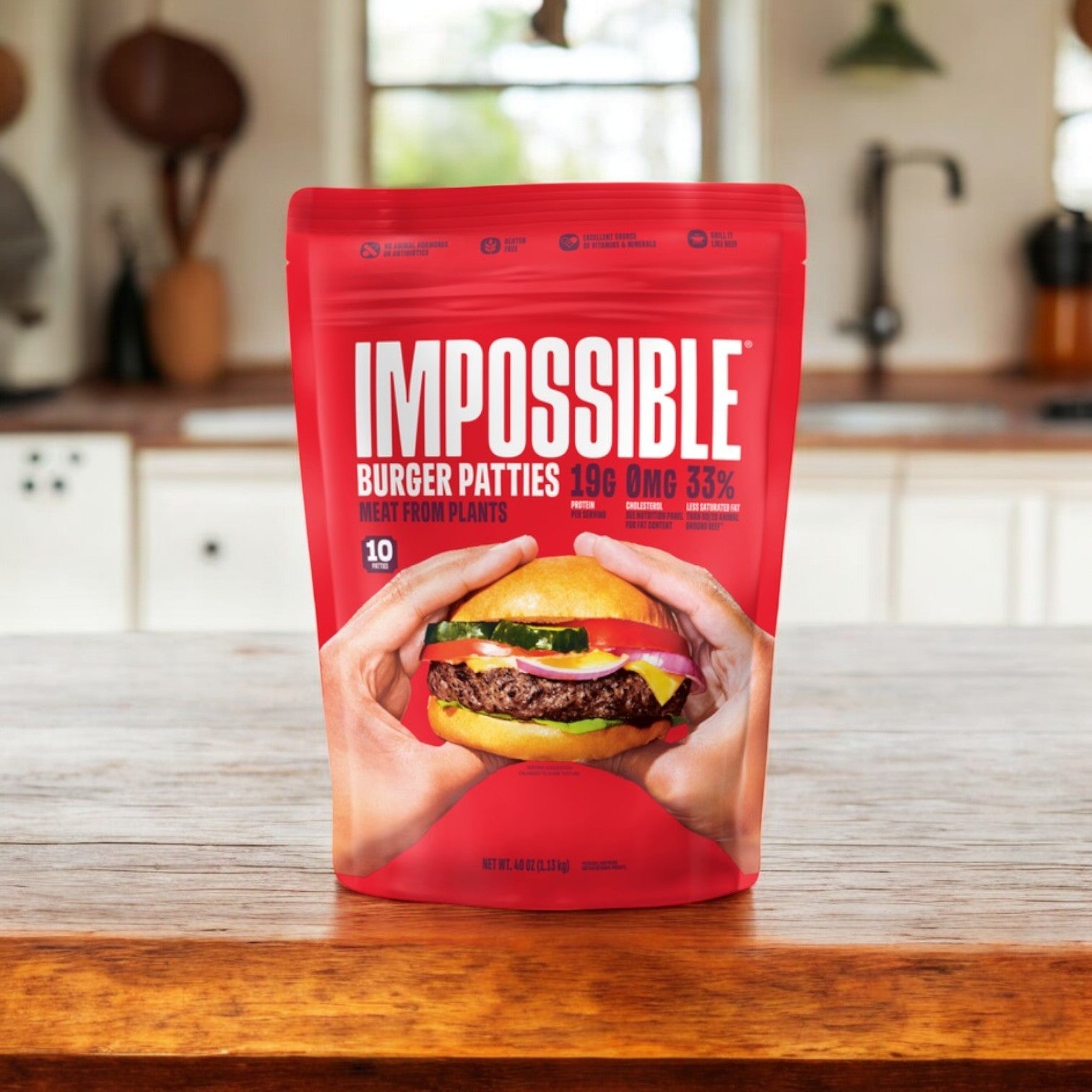 A red package of Impossible Foods' Impossible Burger Patties Made From Plants (10 count, 1 pack) rests on a wooden kitchen counter, featuring a burger with lettuce, tomato, and cheese on the packaging. Kitchen utensils and windows create the backdrop.