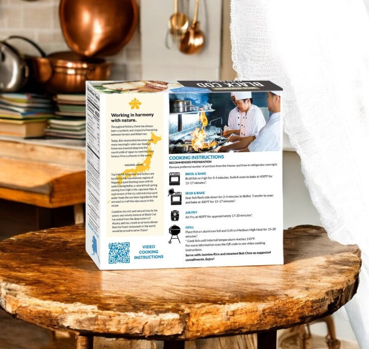On a wooden kitchen table, the Alaskan Leader Seafoods Black Cod, Miso Marinated, 30 oz package displays cooking instructions alongside a map graphic and QR code. In the background, copper pots and premium seafood kitchenware add a touch of elegance.