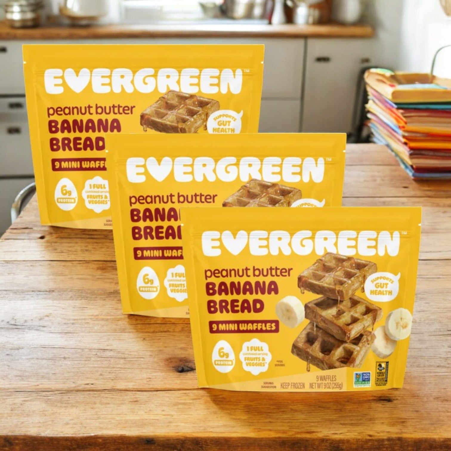 Three packs of Evergreen Peanut Butter Banana Bread Frozen Mini Waffles (9 oz, 3 count) are displayed on a wooden table in a kitchen, perfect for a nutritious breakfast.